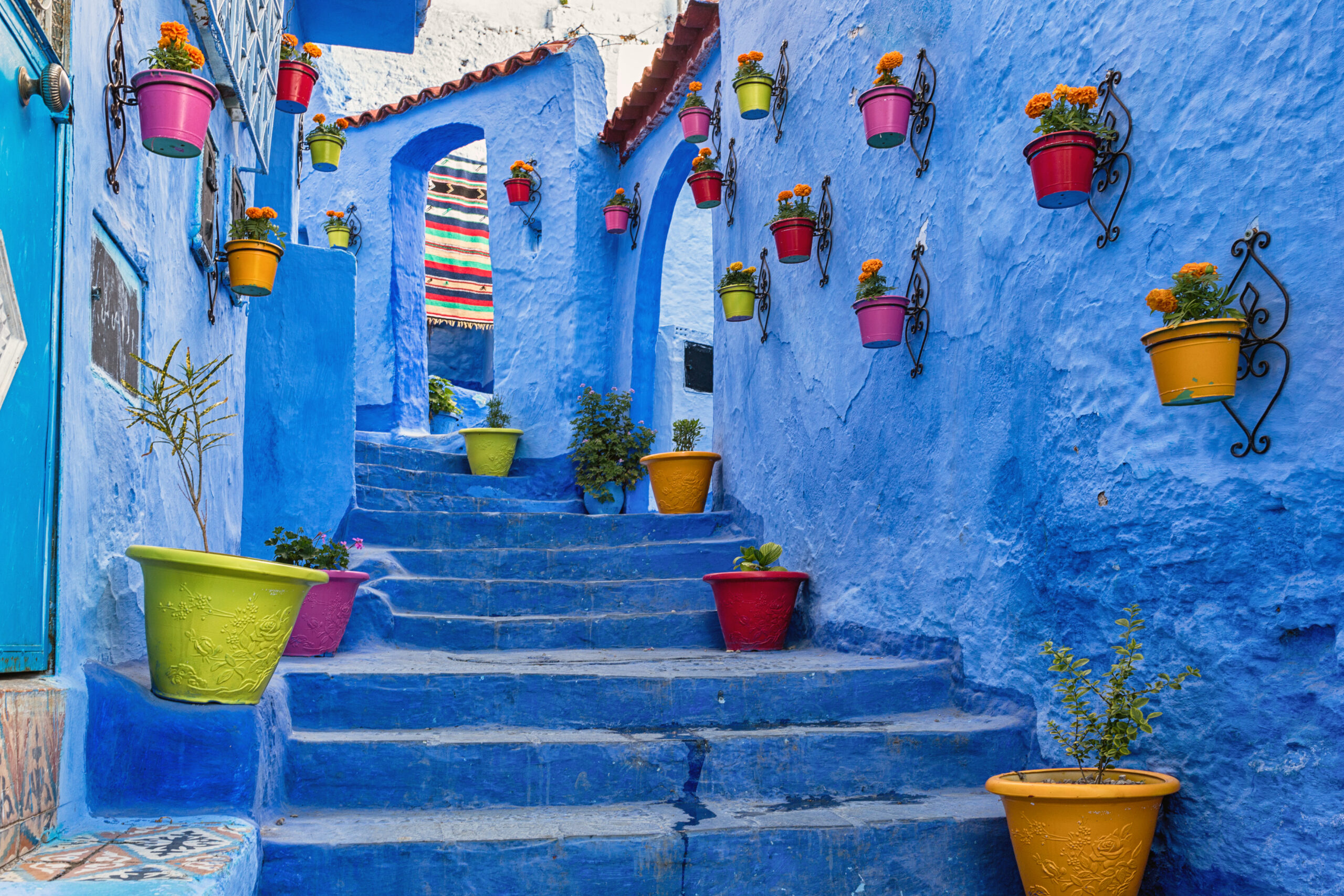 Blue-washed alley with colorful flower pots hanging on walls and along the blue steps.