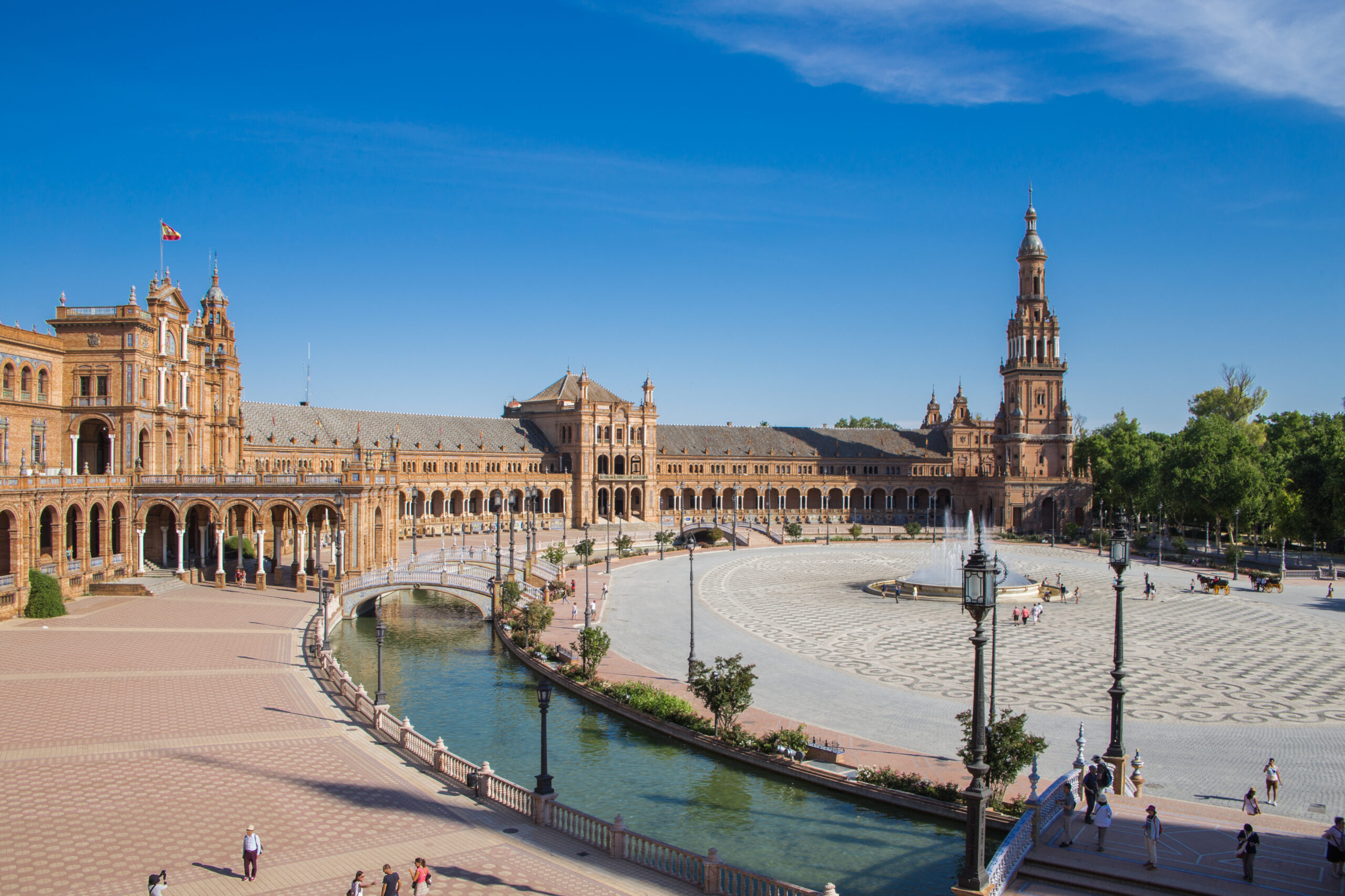 Panoramic view of Plaza de España in Seville, Spain: ornate red-brick arches, a tall tower, and a canal with fountains under a clear blue sky.