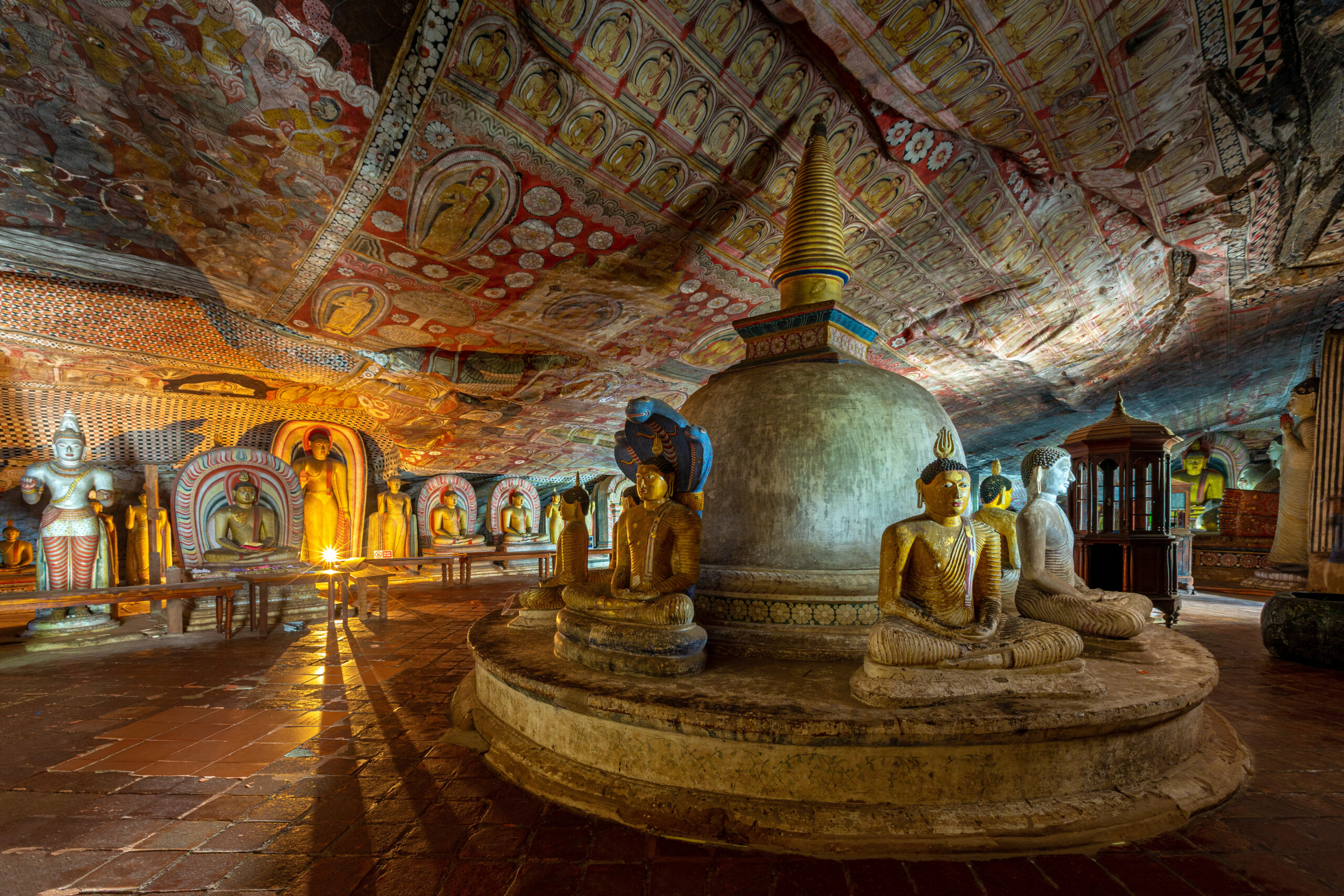 Inside a cave temple filled with colorful murals and a ring of Buddha statues around a central stupa.