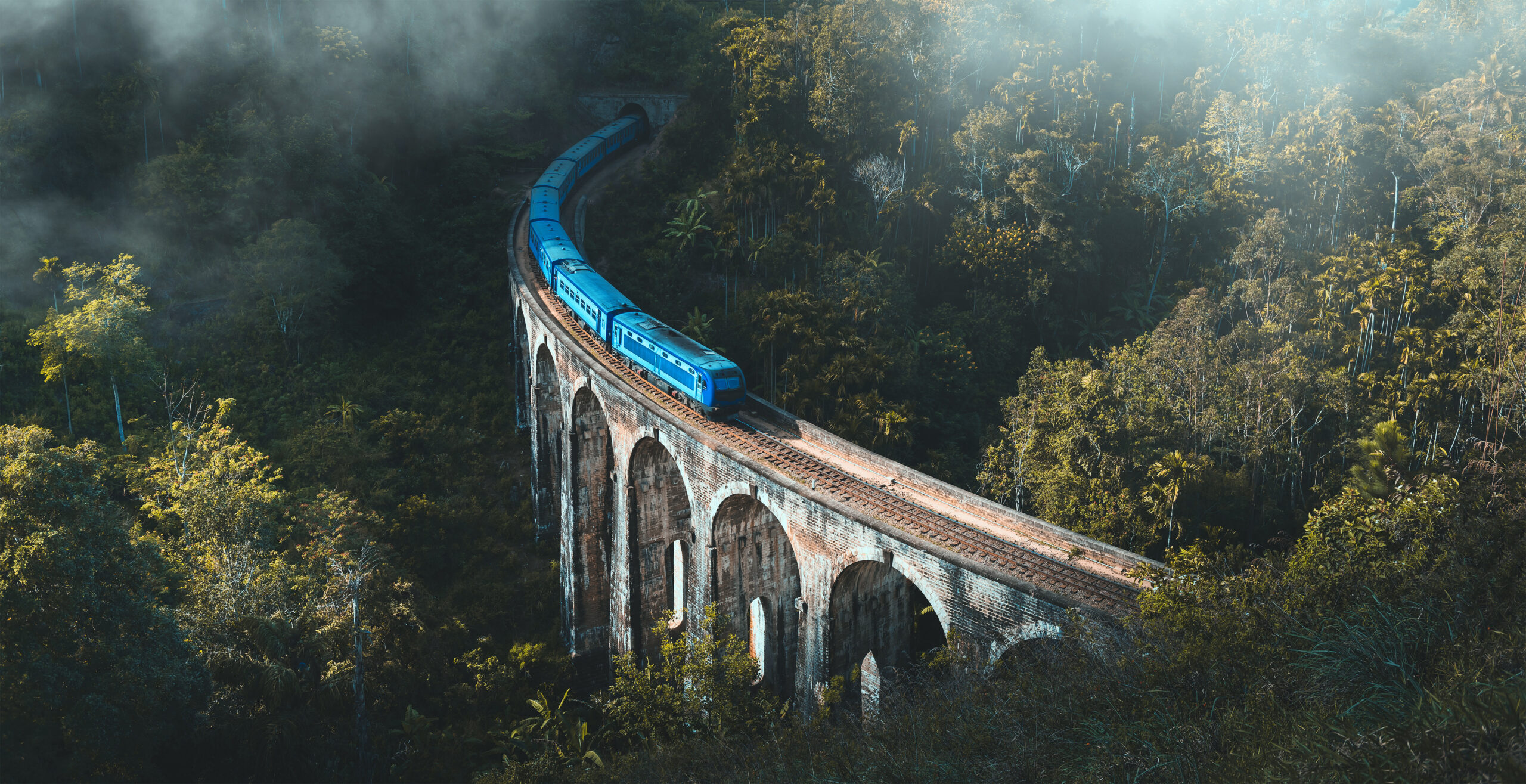 Blue passenger train curves along a brick arch viaduct through dense jungle mist.