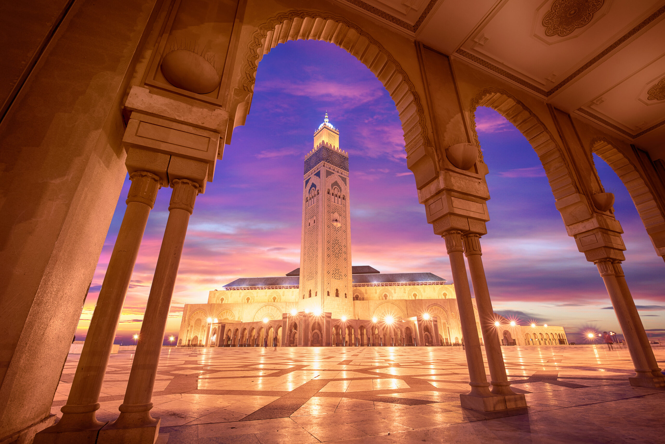 Sunset view of Hassan II Mosque framed by arches and columns, with warm lighting and a blue-purple sky.