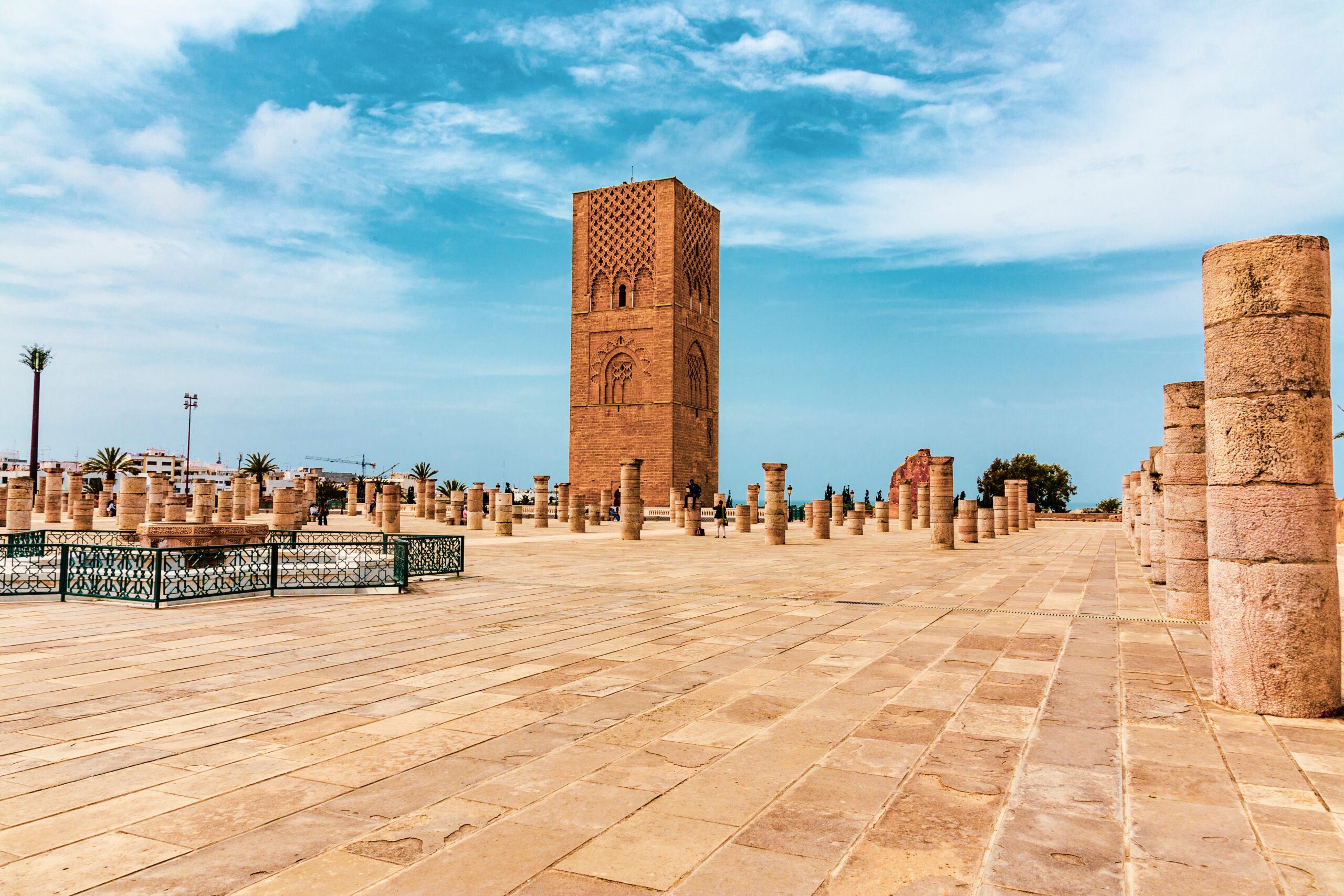 Stone plaza with a tall brick minaret-like tower and rows of columns under a blue sky.