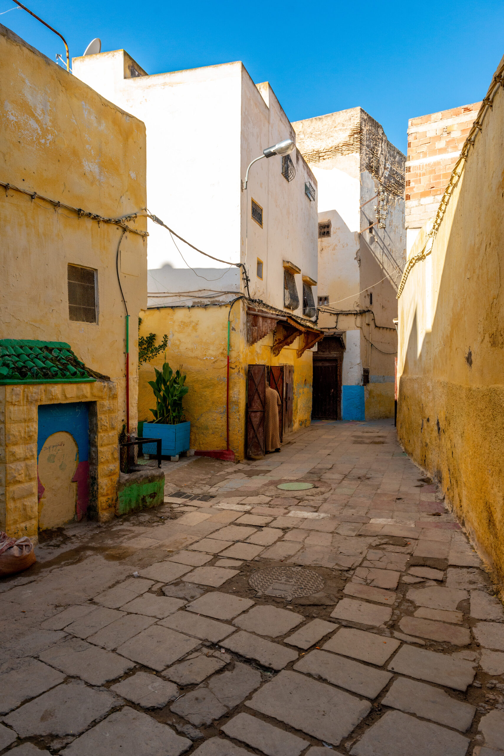 Narrow sunlit alley lined with yellow and white stucco buildings, blue sky above, and a stone-paved path with plants in blue planters on the left.