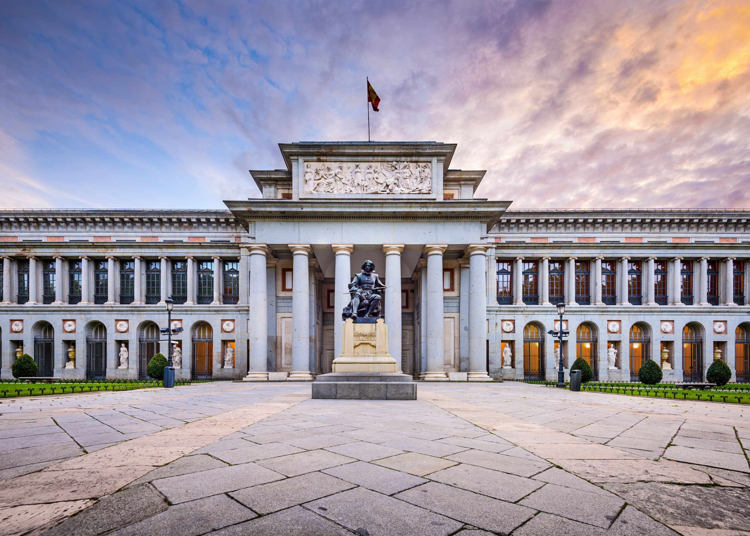 Front view of a neoclassical building with a statue in the courtyard and a flag atop the central pediment against a cloudy sky.