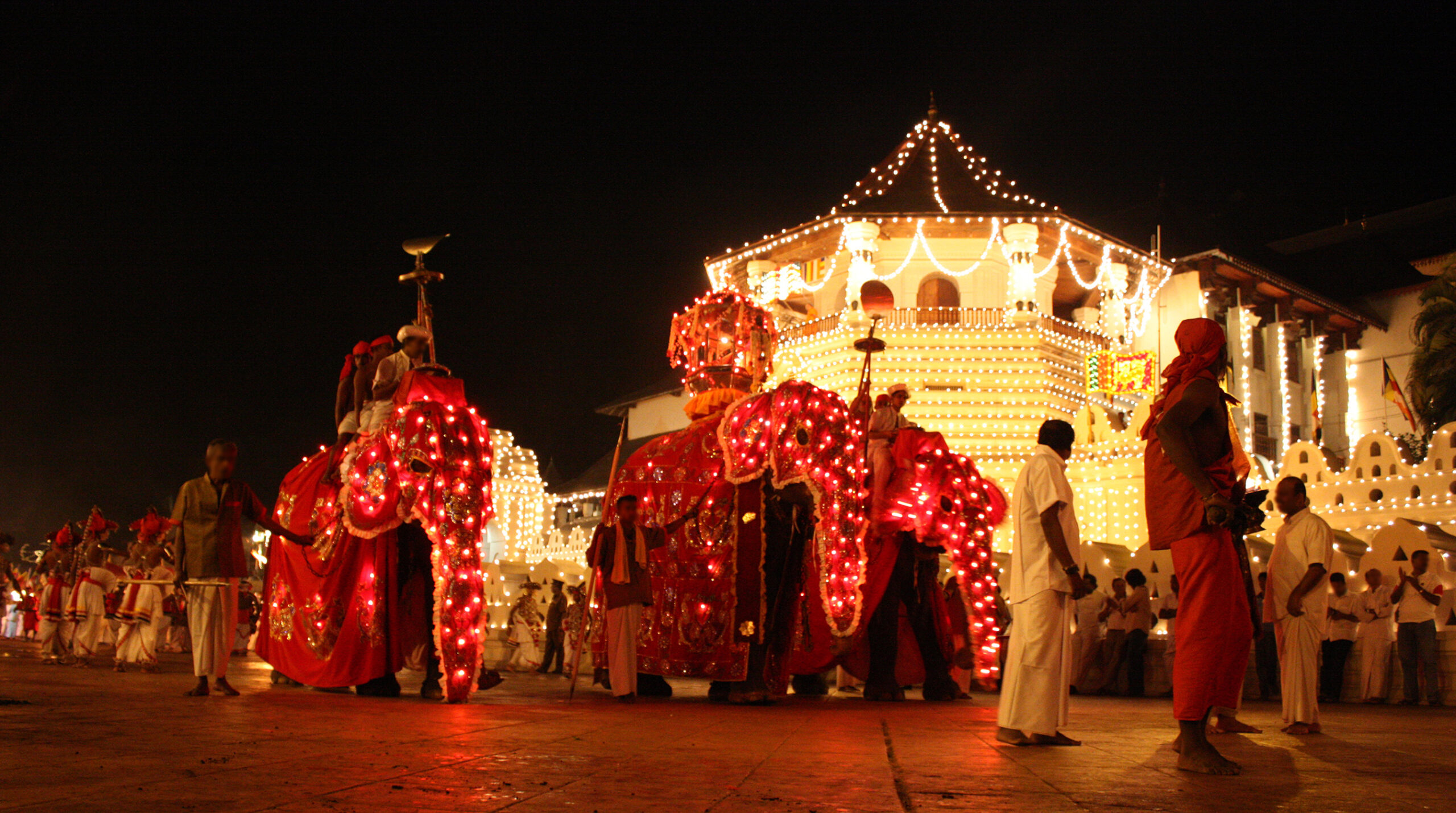 Night parade with illuminated float and performers in traditional clothing; string lights decorate a large, ornate building in the background.