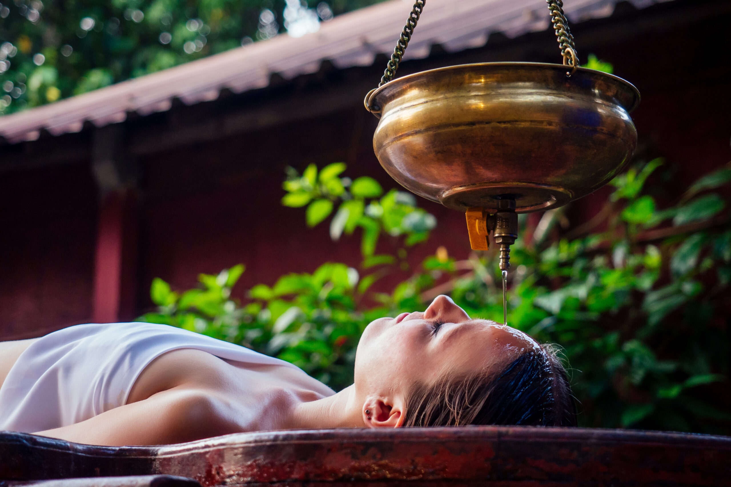 Person lying in a metal tub as a copper vessel pours water over their face, in a spa-like outdoor setting.