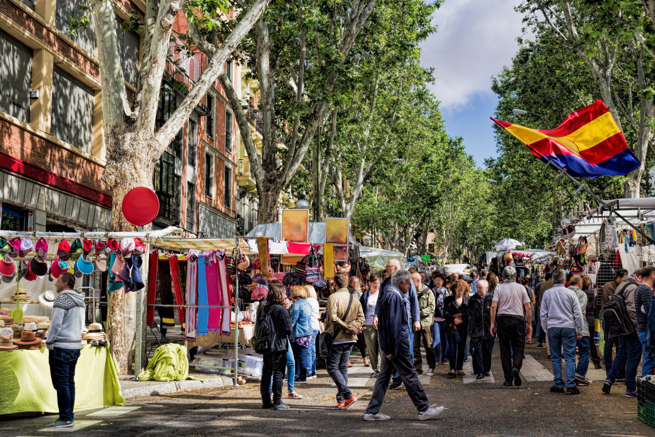 Busy outdoor street market with stalls selling hats and clothing under tall trees.