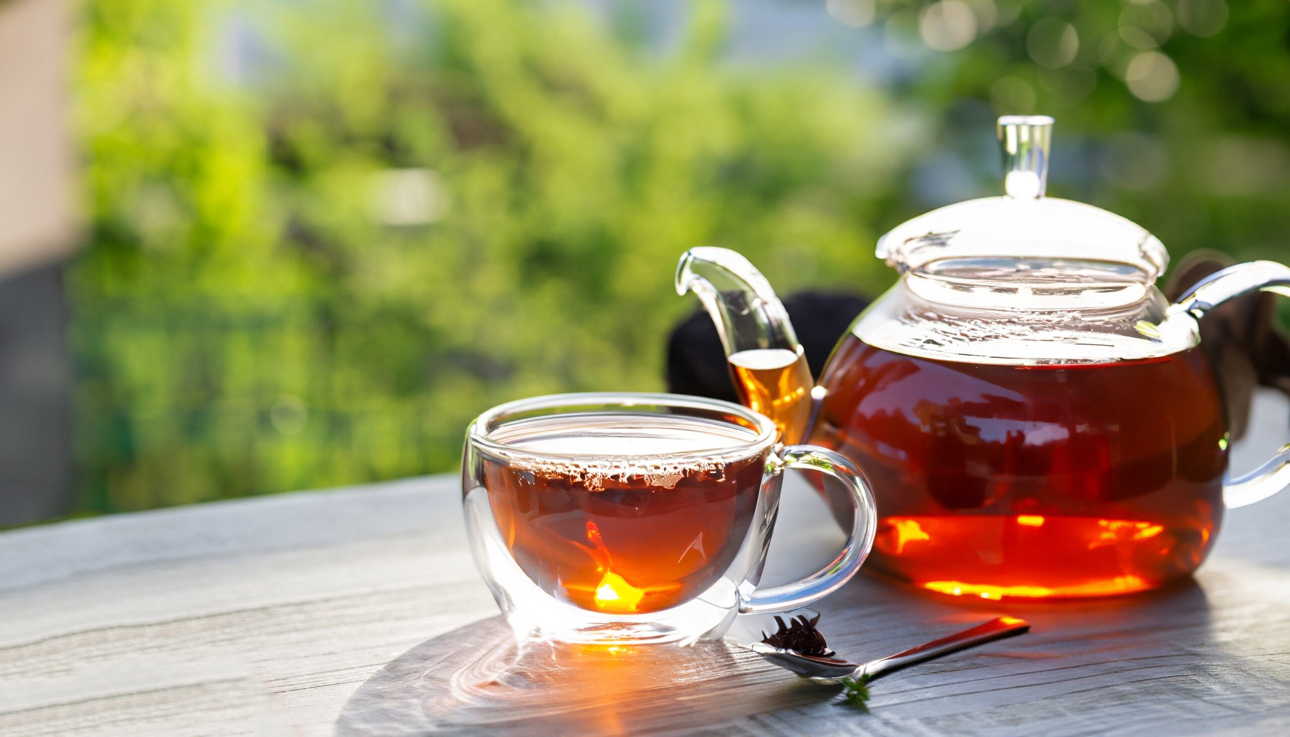 Glass teapot and two glass cups filled with tea on a wooden table outdoors with a green blurred background