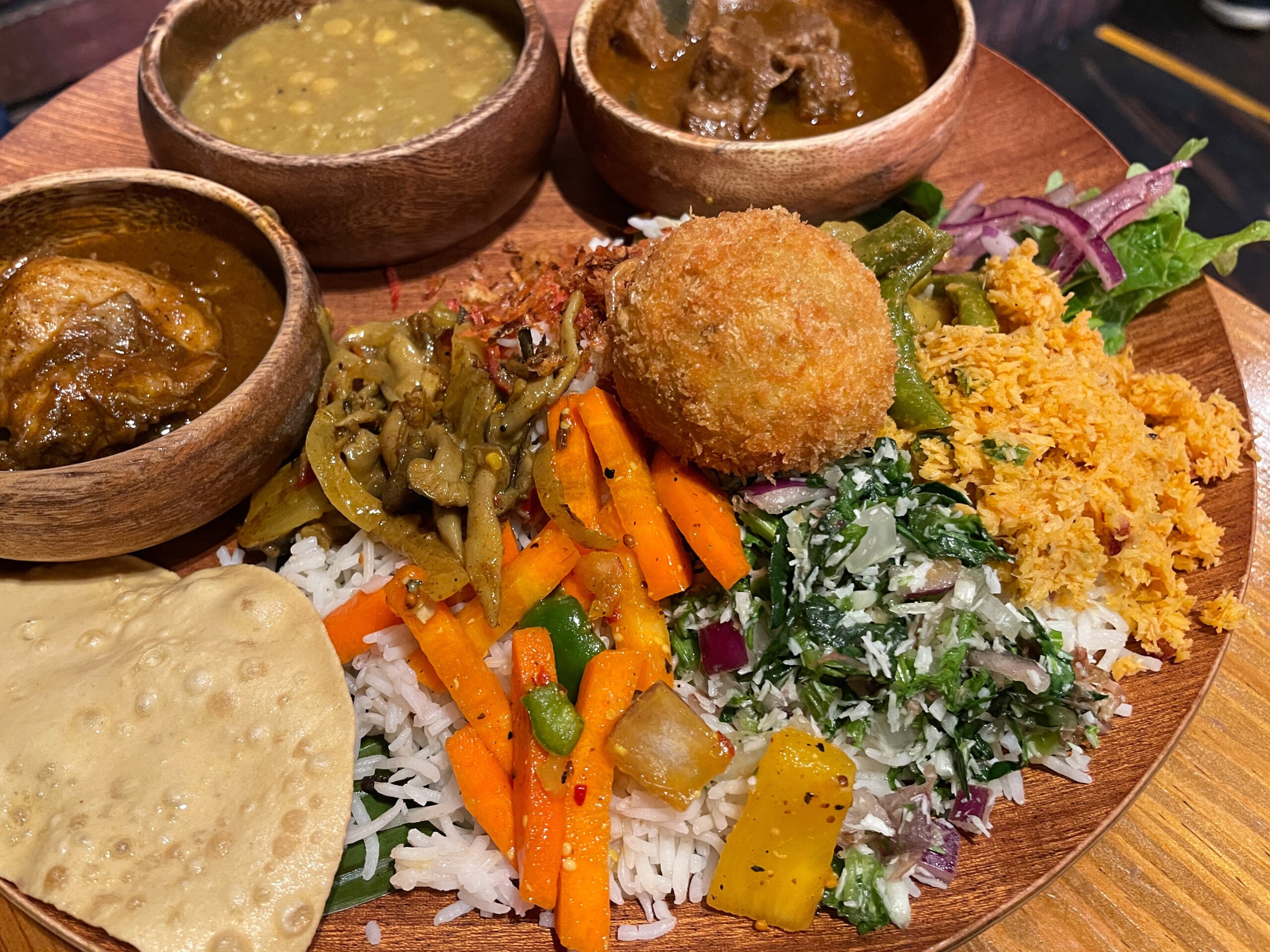 A wooden platter with rice, three curries in bowls, fried croquette, mixed vegetables, papad and greens on the side