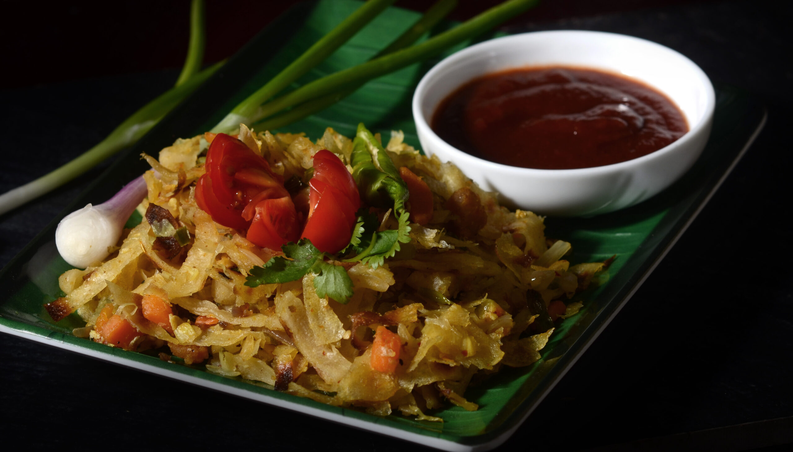 Crispy fried rice with vegetables, tomatoes, and cilantro on a banana-leaf plate, with a white bowl of red dipping sauce