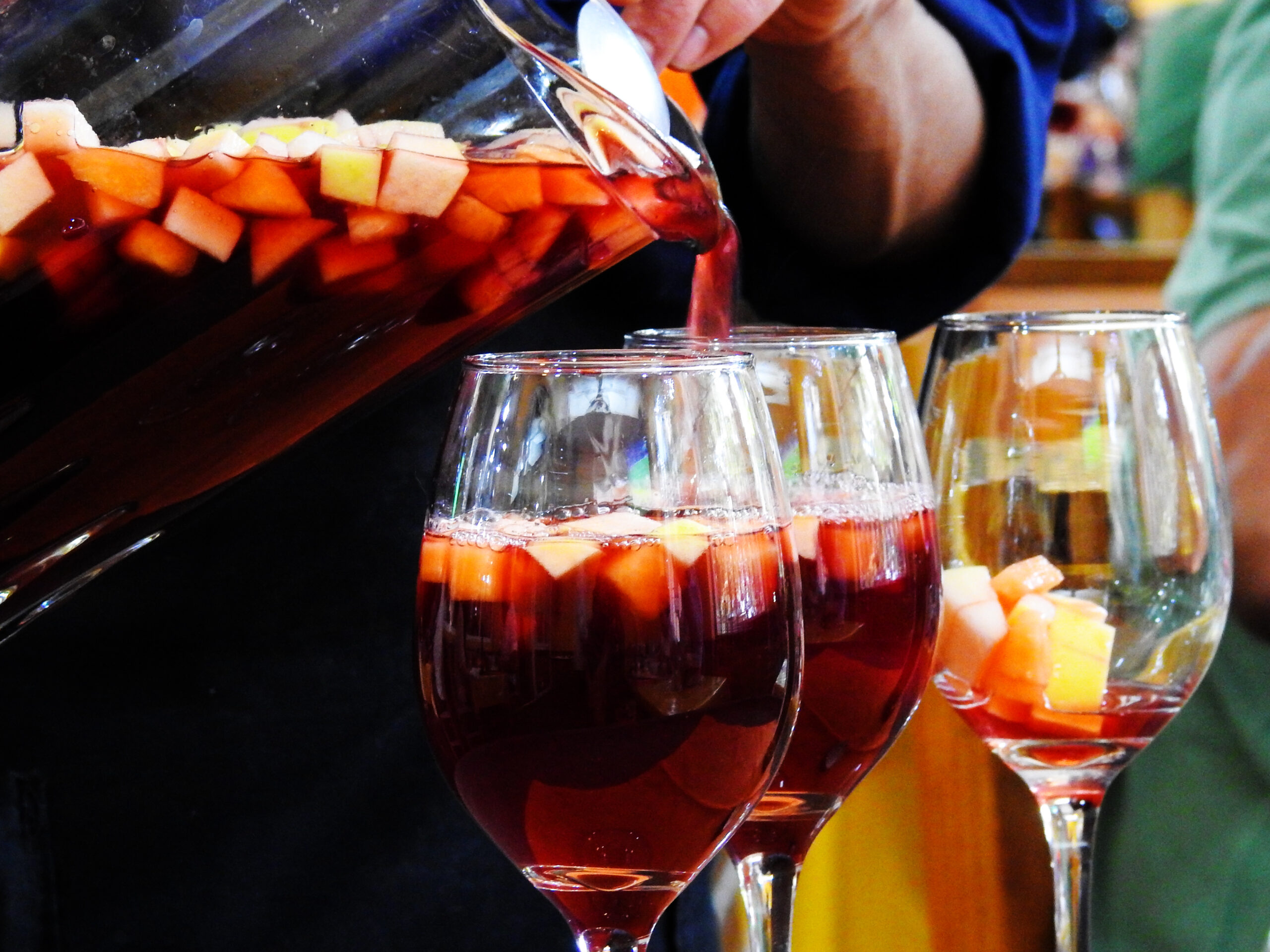 Pouring red sangria with chopped fruit into stemmed glasses from a pitcher.