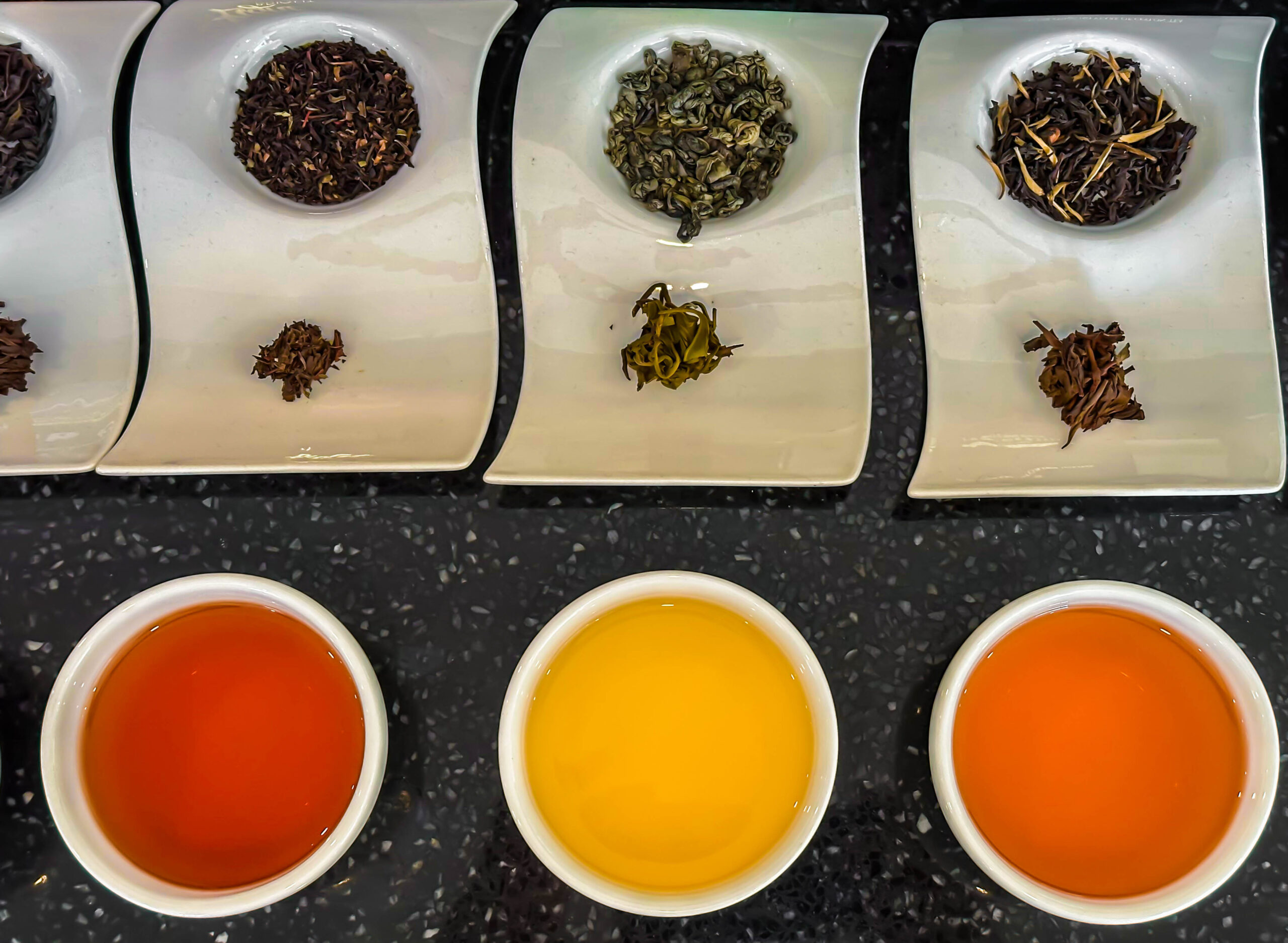 Tea sampler flight: five cups of hot tea ranging from orange to yellow on a dark countertop, with piles of tea leaves on white plates above each cup.