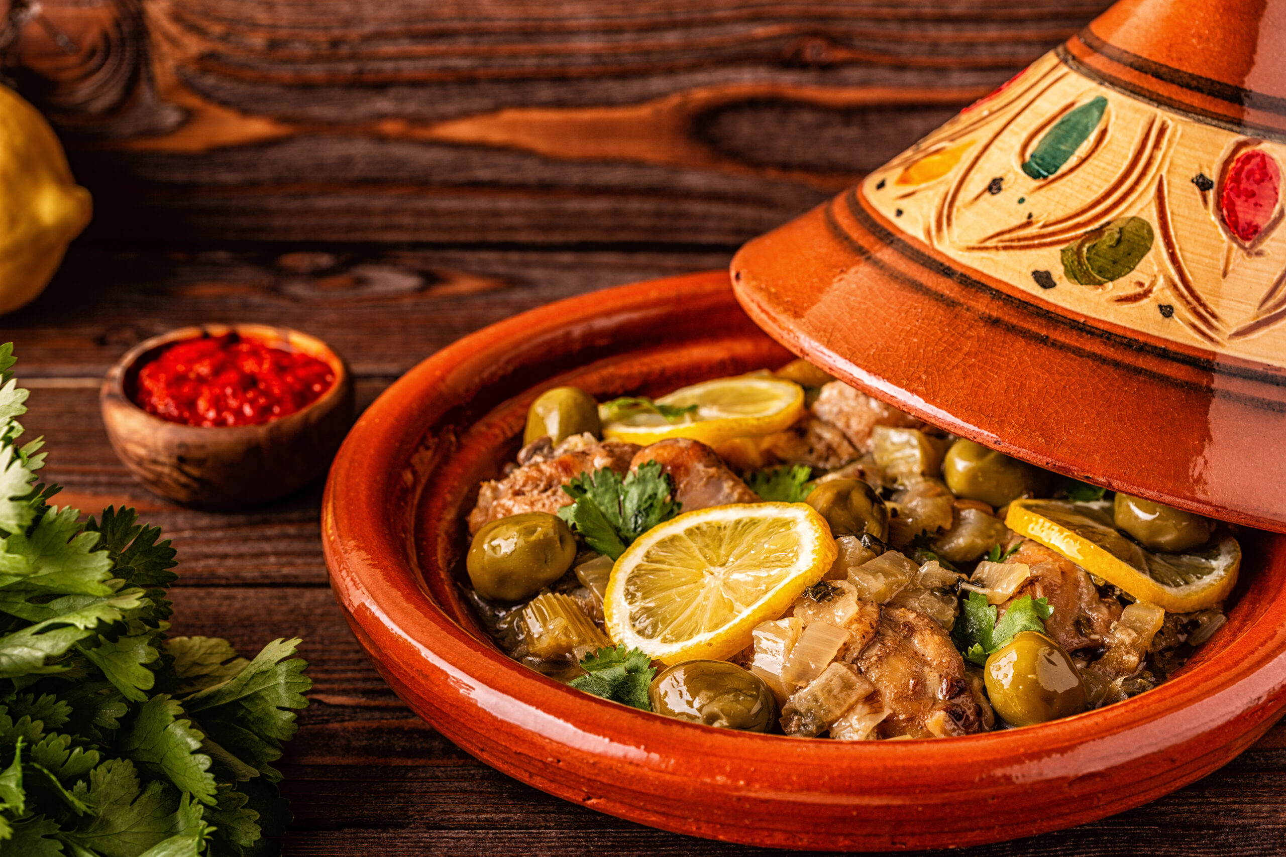 Clay tagine half-open to reveal meat, green olives, and lemon slices, garnished with parsley on a wooden table with a side of red sauce nearby