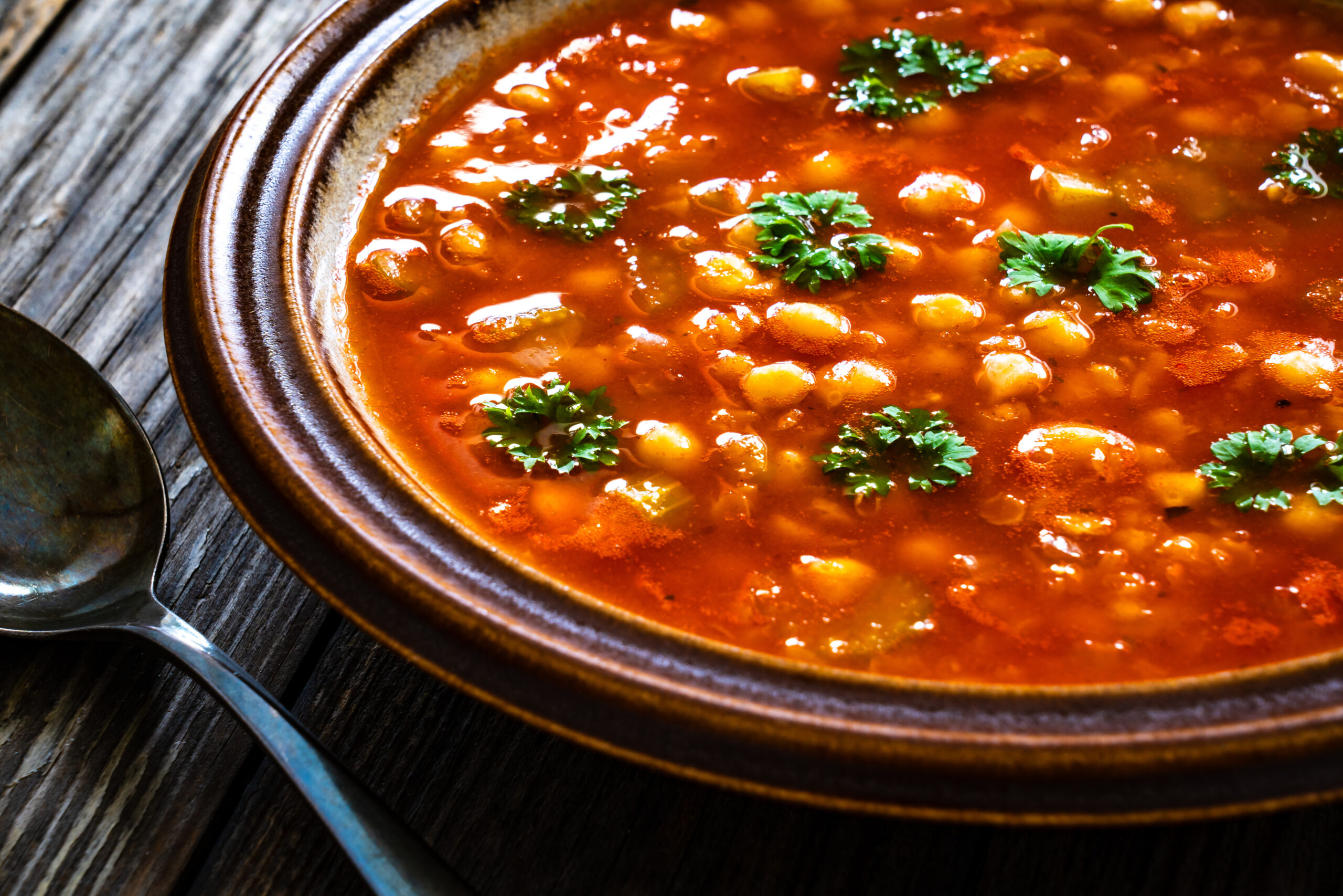 Close-up of a red chickpea soup garnished with parsley in a brown ceramic bowl on a wooden table.
