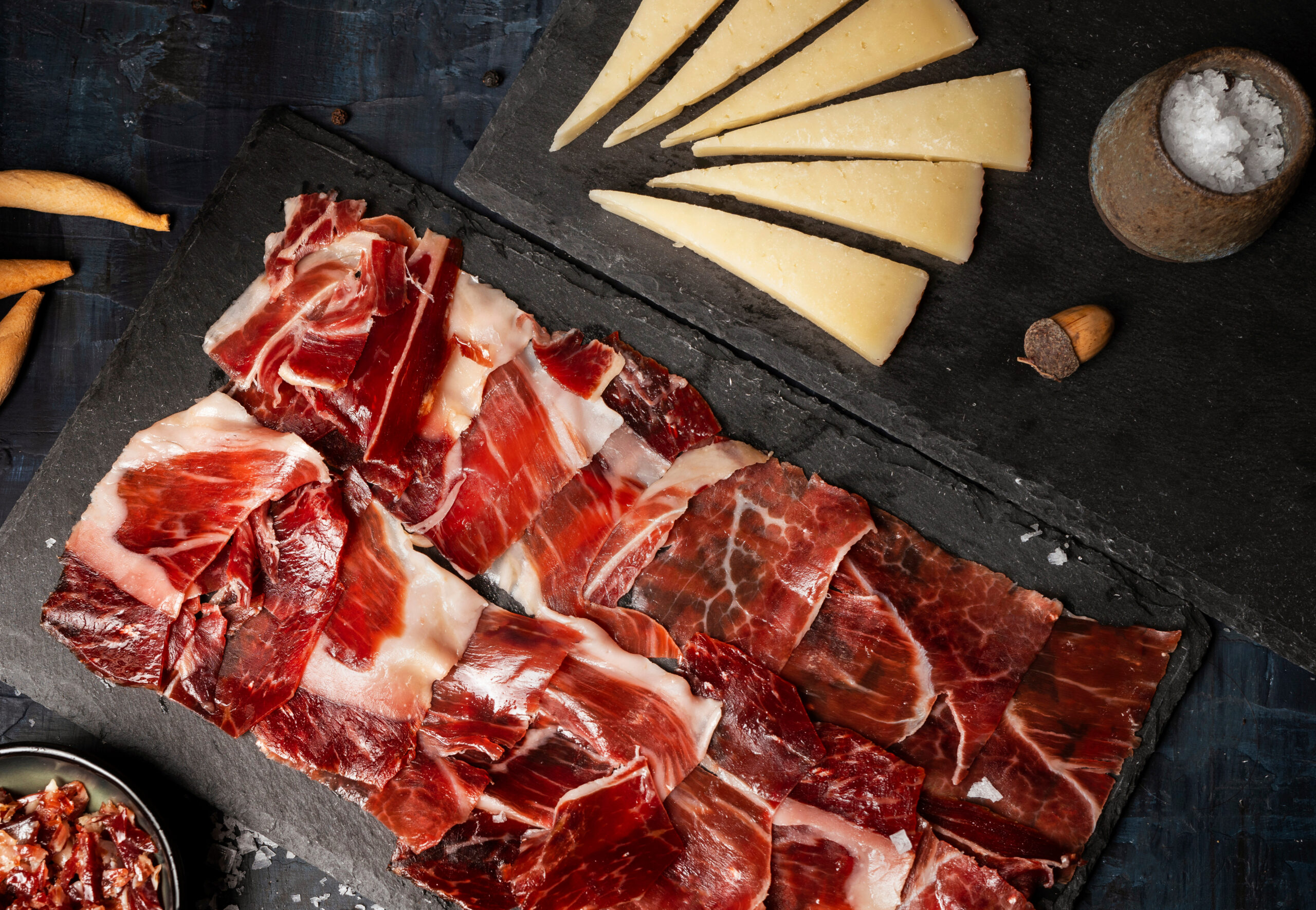 Charcuterie board with thinly sliced cured meat and wedge-shaped cheese on a slate surface, salt in a rustic bowl to the side.