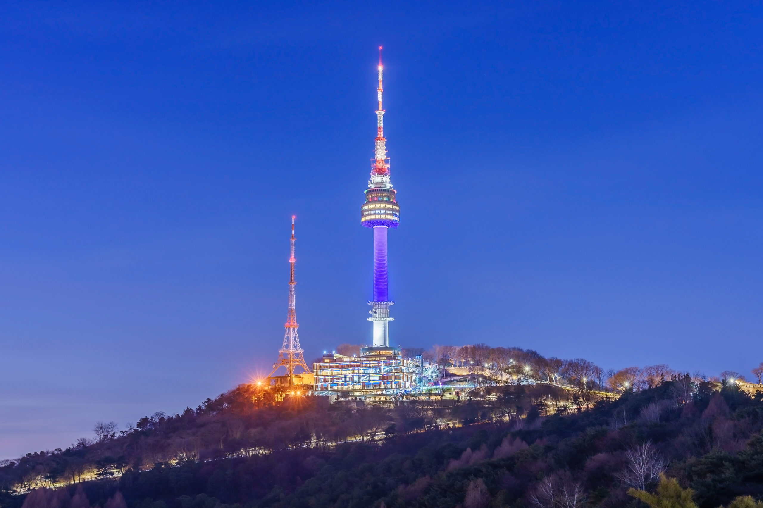Tall illuminated communications tower on a hill at dusk, with a smaller tower to the left and a lit complex at the base.