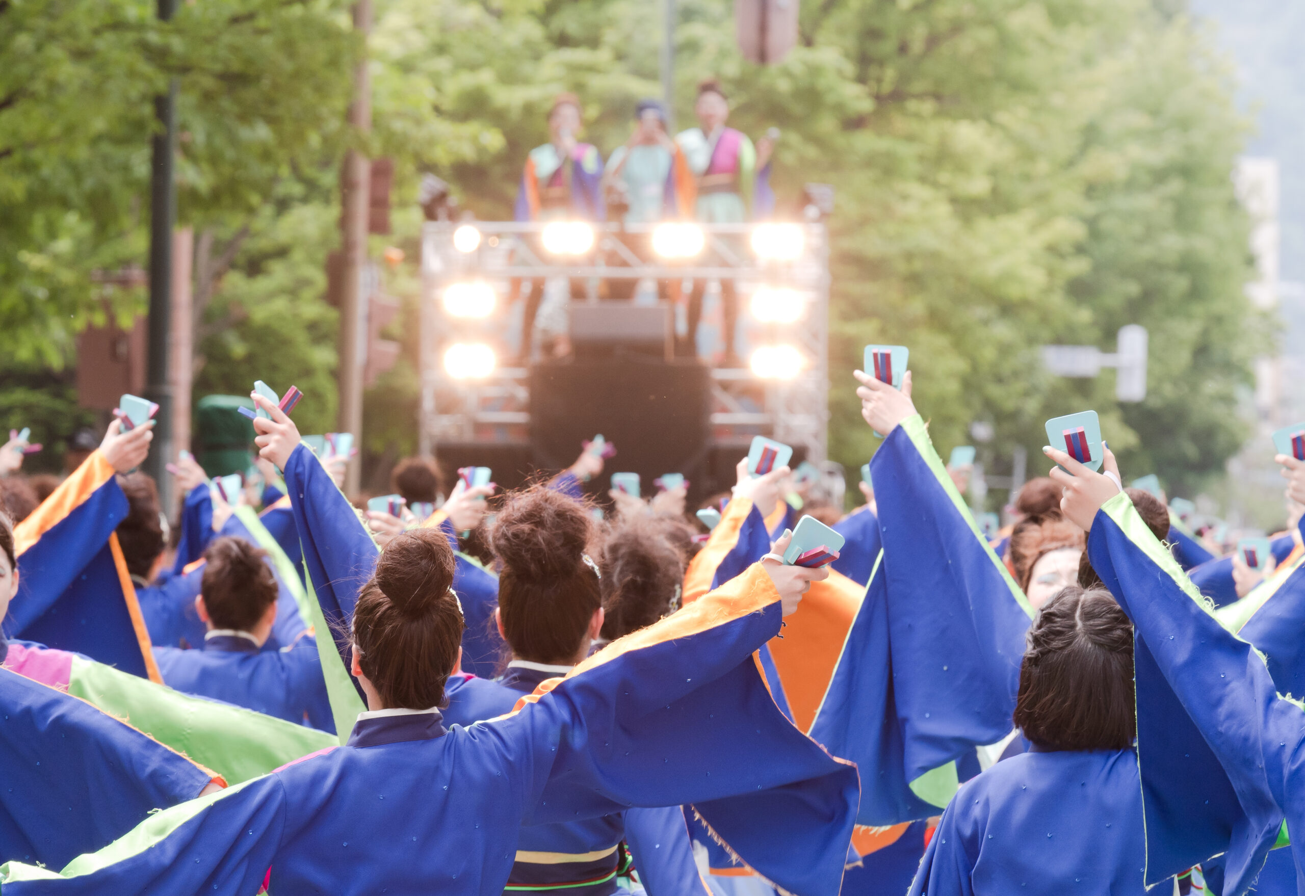 Parade crowd in blue robes, holding up phones, facing a brightly lit float with people on top in colorful outfits.
