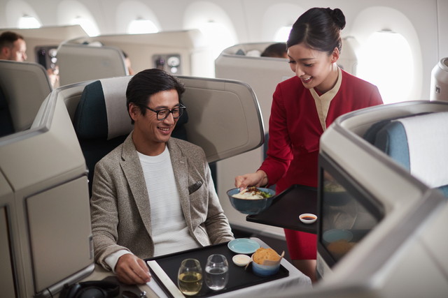 Flight attendant in a red uniform serves a passenger in business class; man smiles at his tray of food and wine.