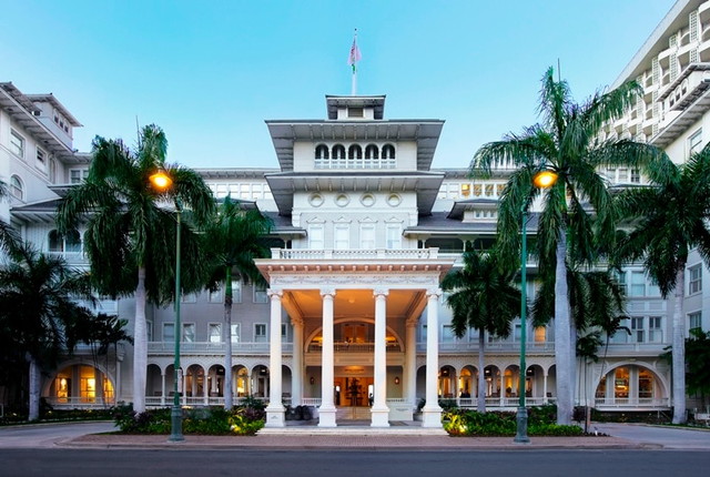 Front view of a grand white hotel with a columned portico and palm trees, illuminated at dusk.
