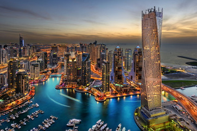 Aerial view of a harbor city at sunset with illuminated high-rise buildings and a marina along winding blue water.