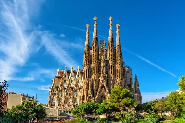 Exterior view of the Sagrada Família cathedral in Barcelona, showing its tall ornate spires and detailed stonework against a bright blue sky with greenery in the foreground.