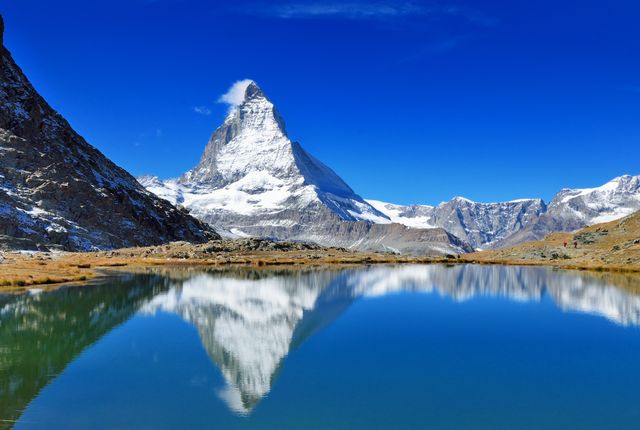 Snowy, pyramid-shaped mountain reflected in a calm alpine lake beneath a bright blue sky.