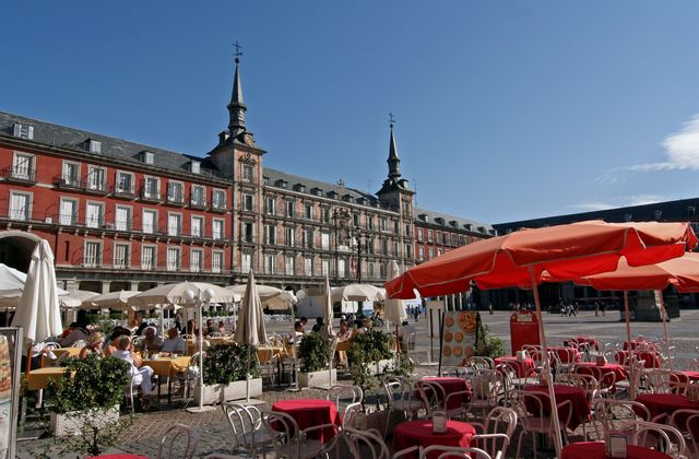 Cafe terrace in a sunny square with red tables and white umbrellas, facing a grand historic building with two spires.