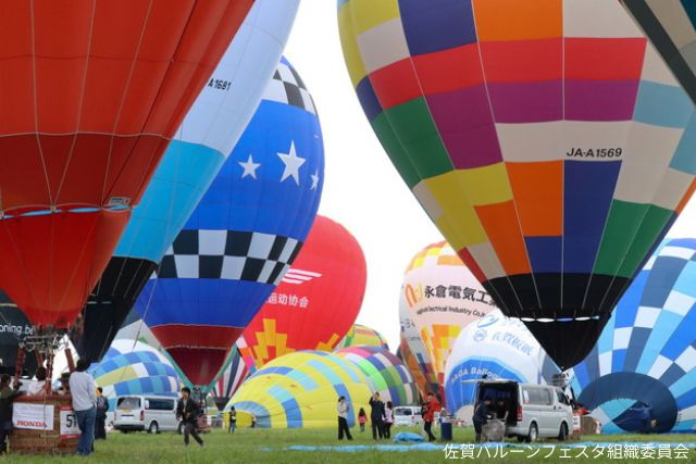 Colorful hot air balloons being inflated on a grassy field, with people and vans nearby under a bright sky.