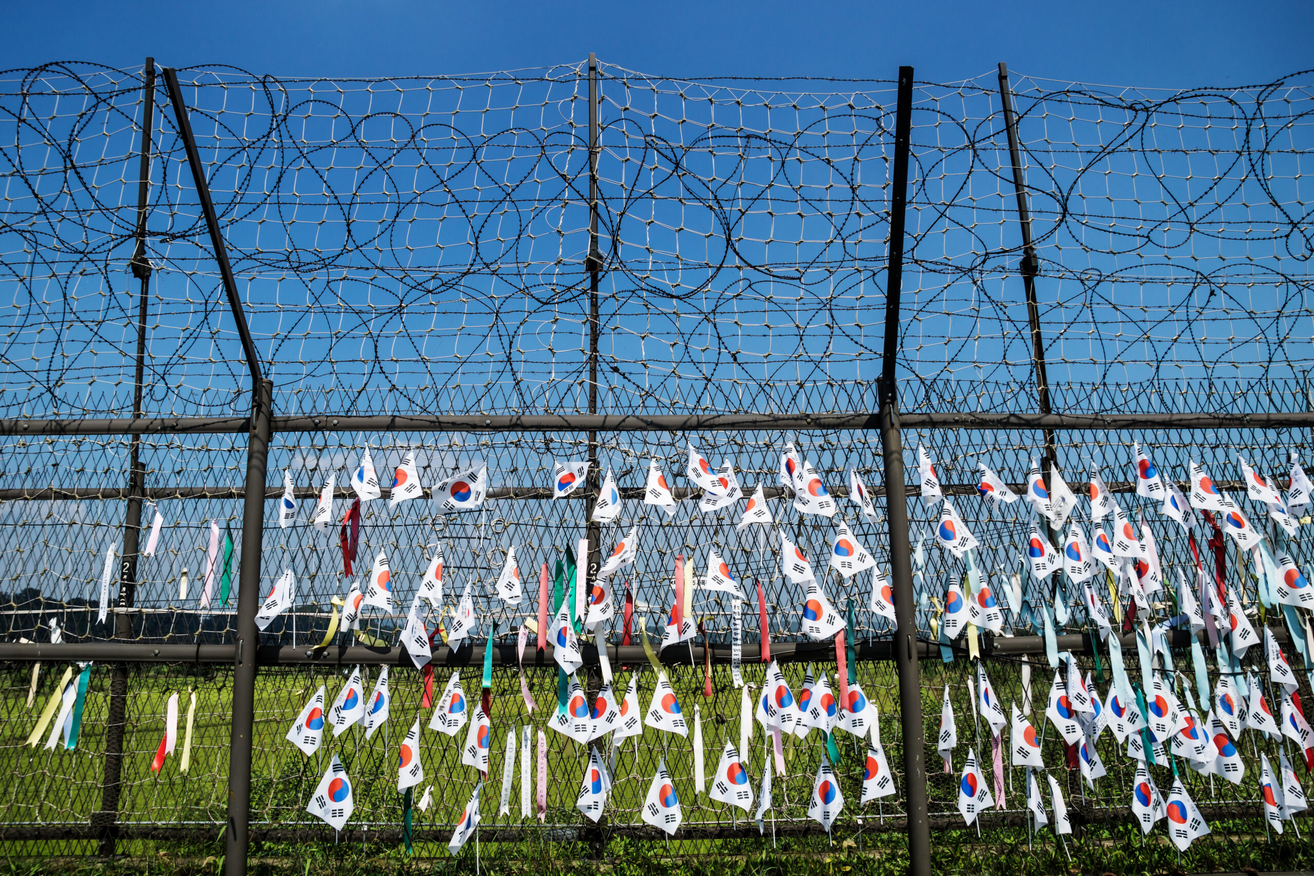 Barbed-wire fence with many small South Korean flags fluttering against a clear blue sky.