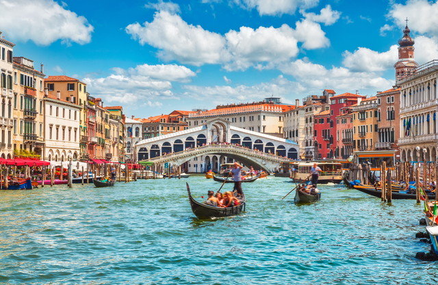 Gondolas glide on the Grand Canal in Venice under the Rialto Bridge, with colorful buildings lining the waterway.