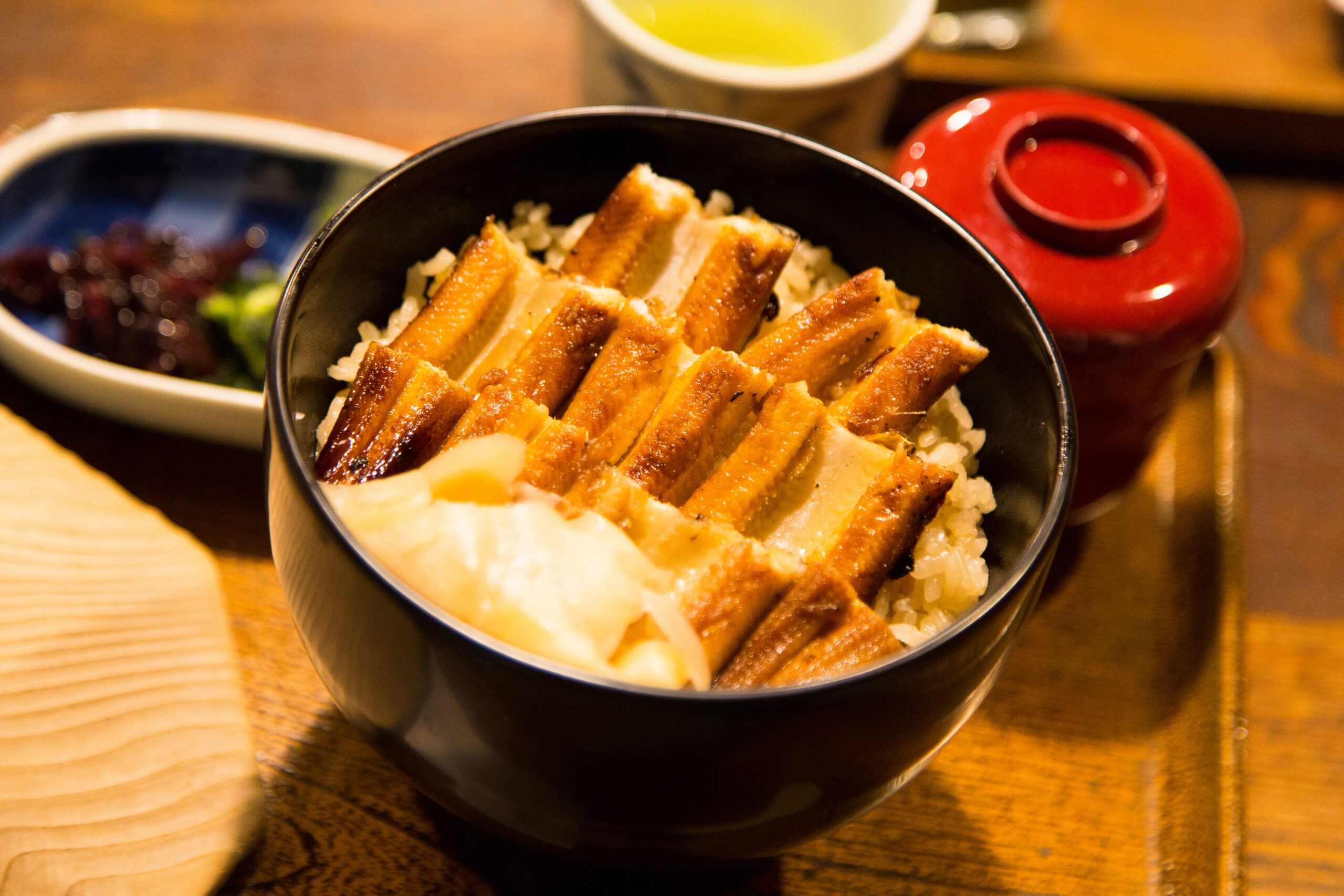 Bowl of grilled eel slices over white rice in a black bowl, on a wooden table.