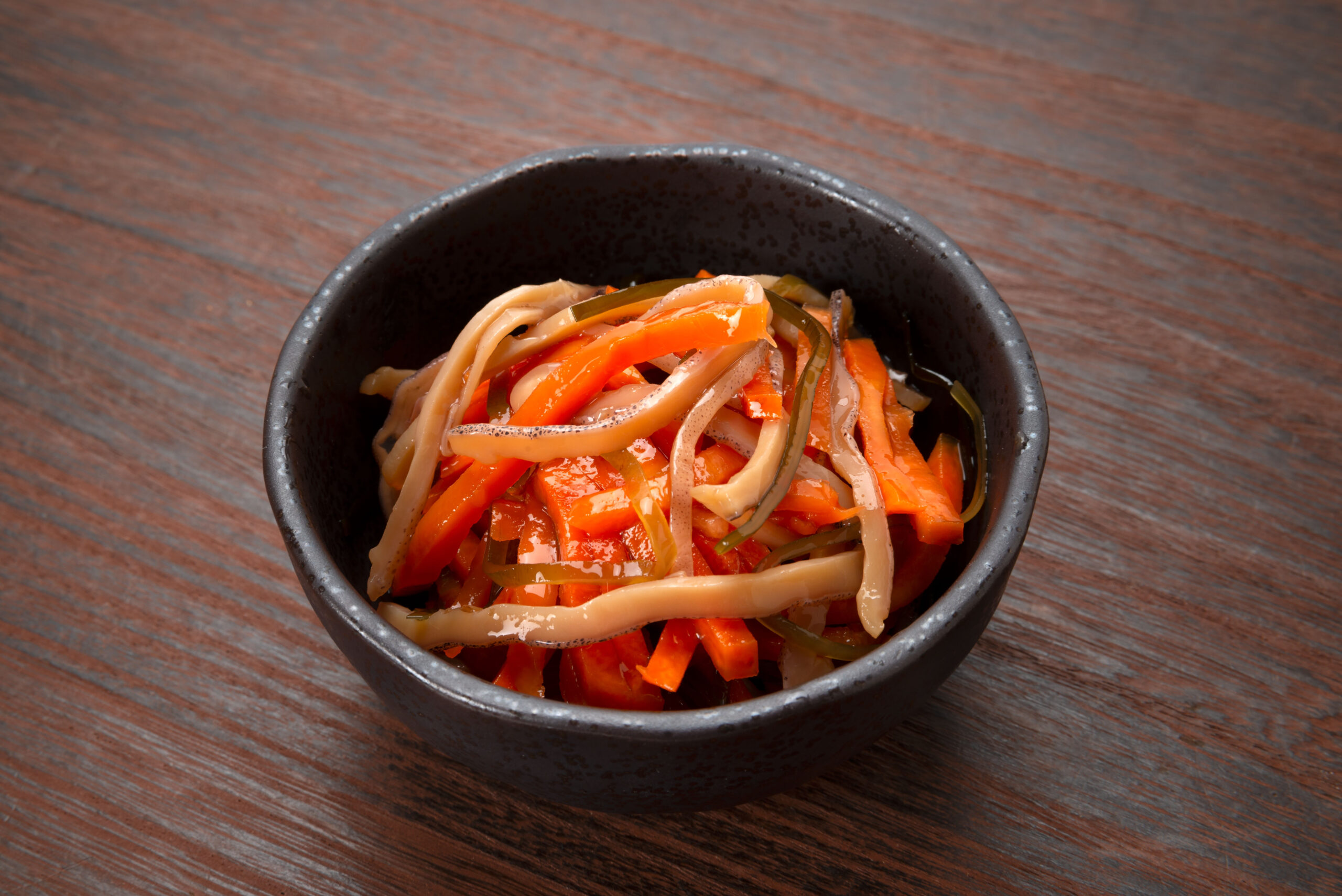 Julienned carrots, bell peppers, and seaweed in a glossy sauce served in a dark ceramic bowl on a wooden table.