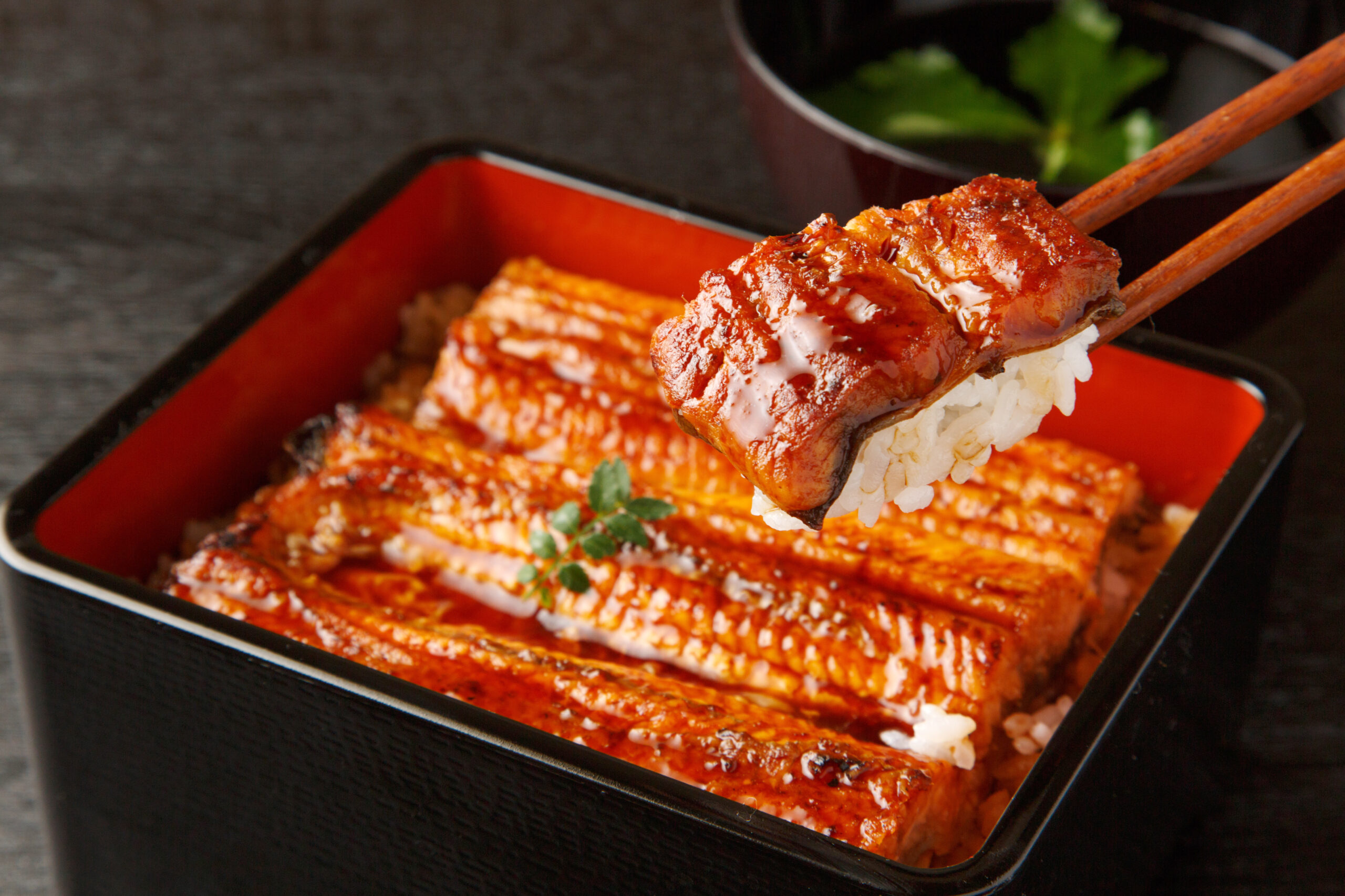 Grilled eel with glaze served over white rice in a traditional black lacquered bento box with chopsticks nearby.