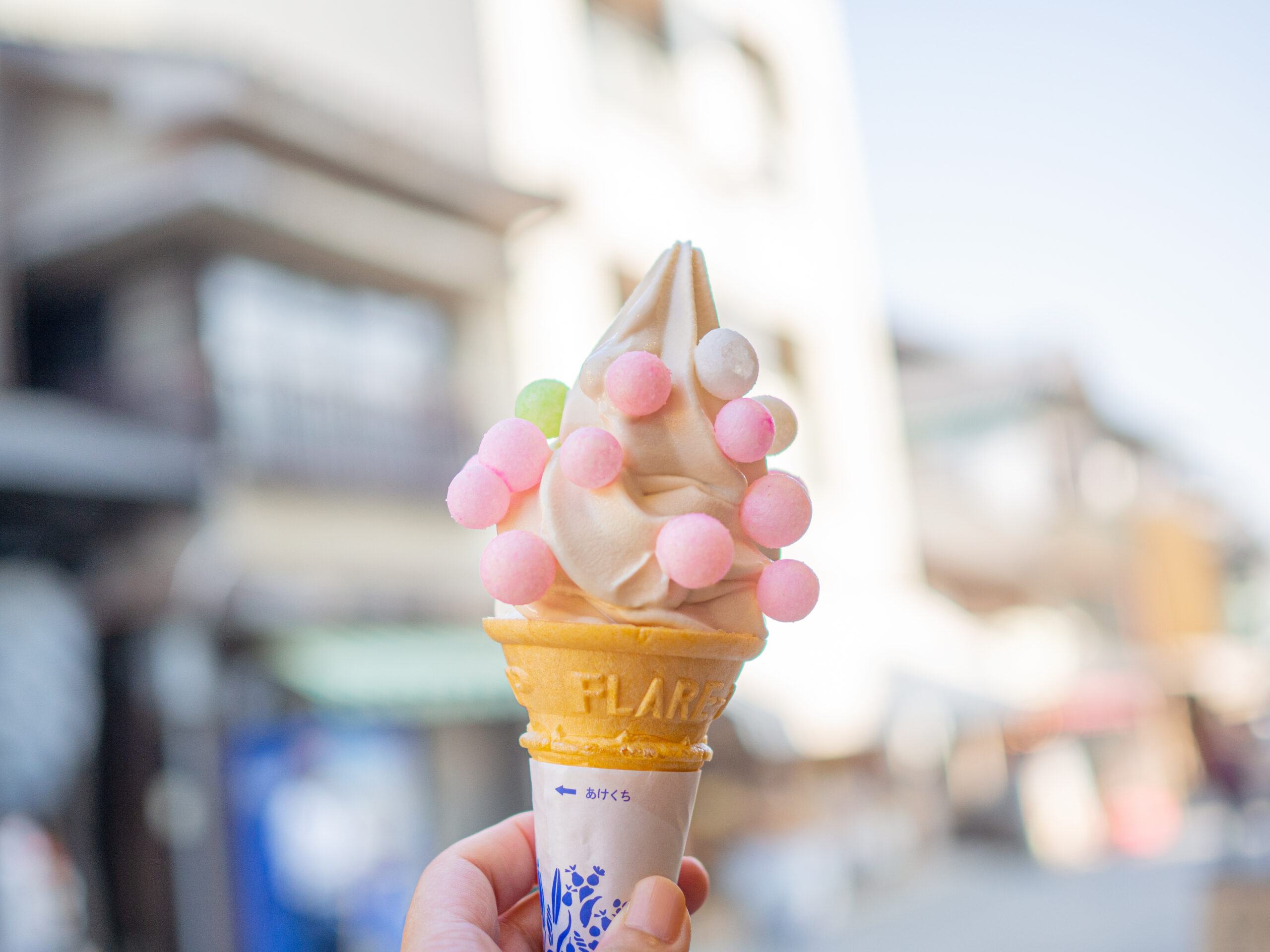 Vanilla soft-serve ice cream cone decorated with pastel pink and white candy beads, held outdoors by a hand.