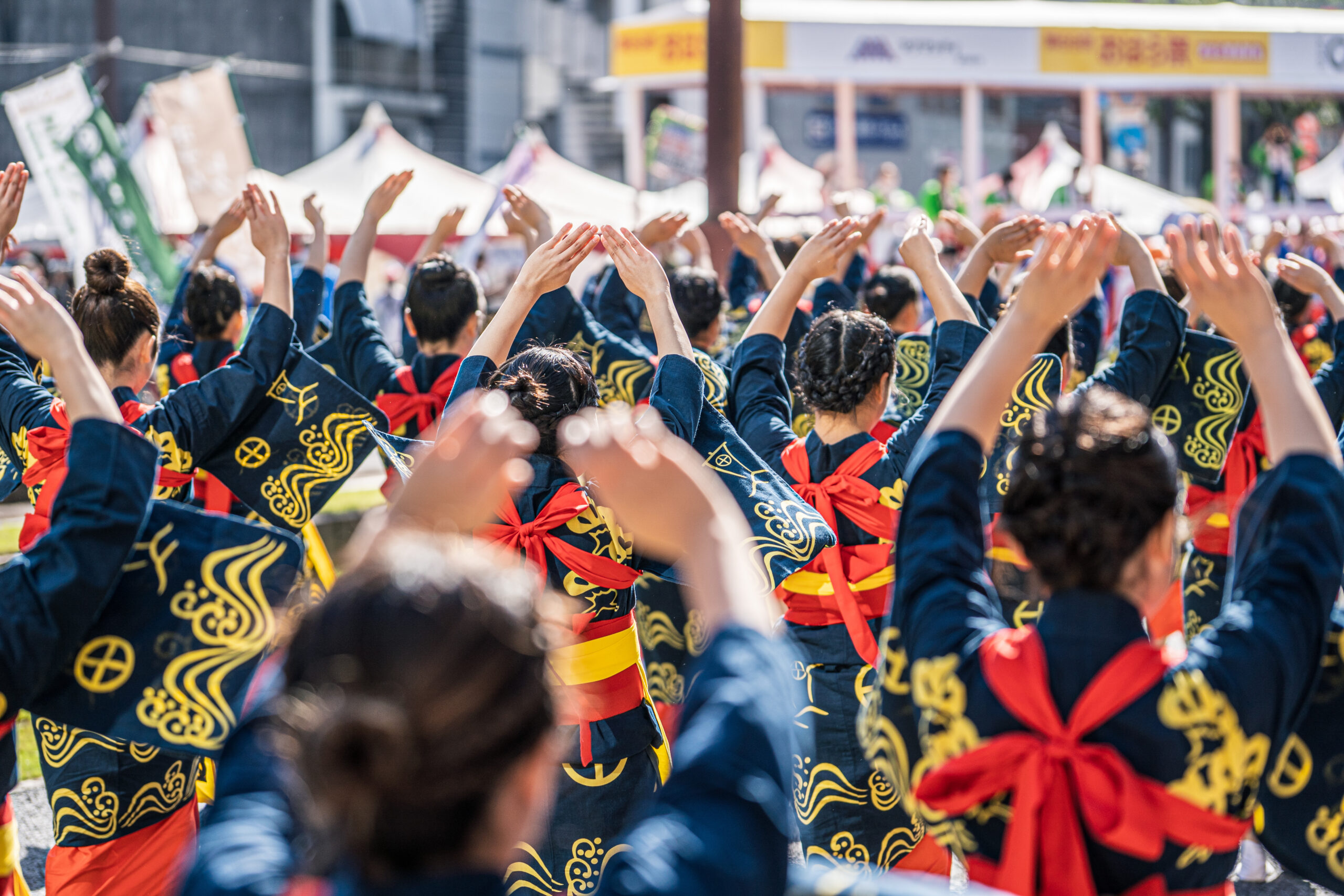 Crowd in blue and gold outfits raises arms in a synchronized dance at an outdoor festival stage far in the background mentions tents and banners.