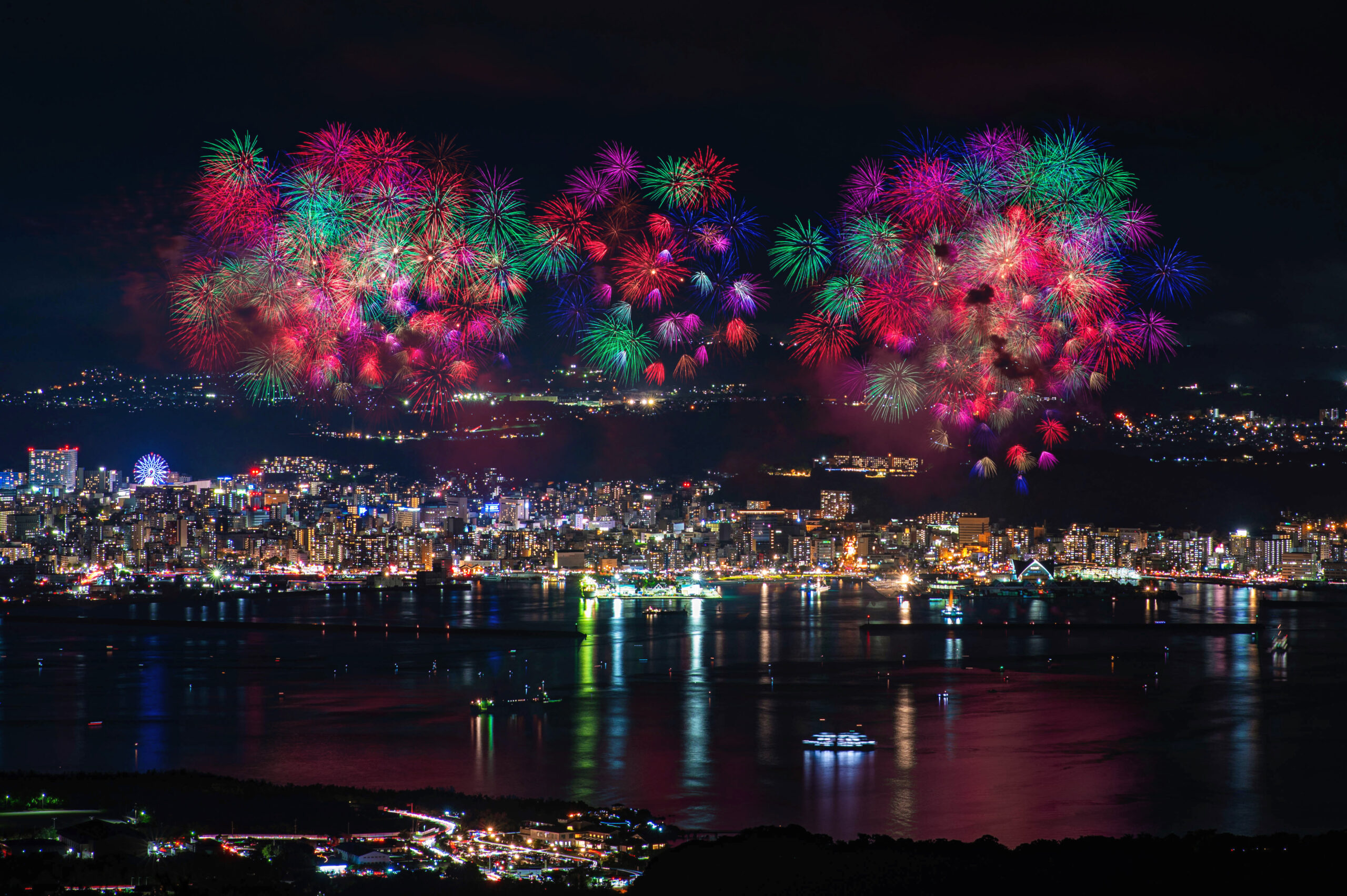 Colorful fireworks explode over a city skyline at night, reflecting on the water below within a harbor setting.