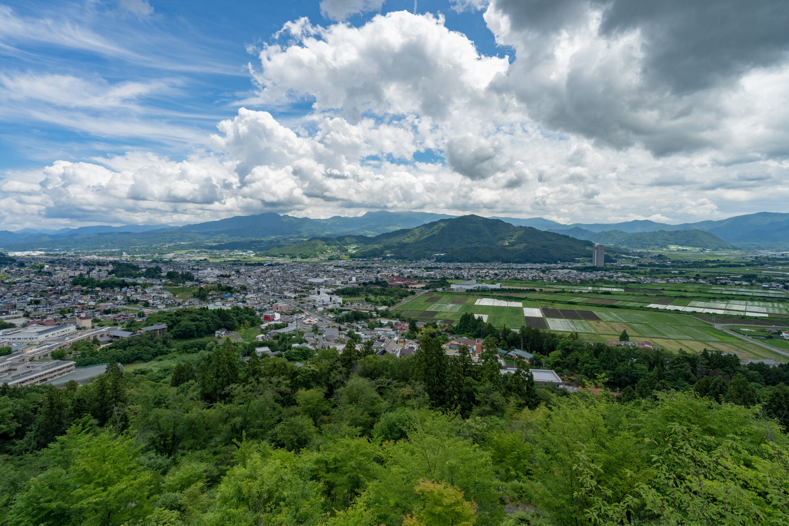 Wide valley view showing a city at the base of green, rolling mountains under a partly cloudy blue sky; patchwork fields in the foreground and trees framing the scene.
