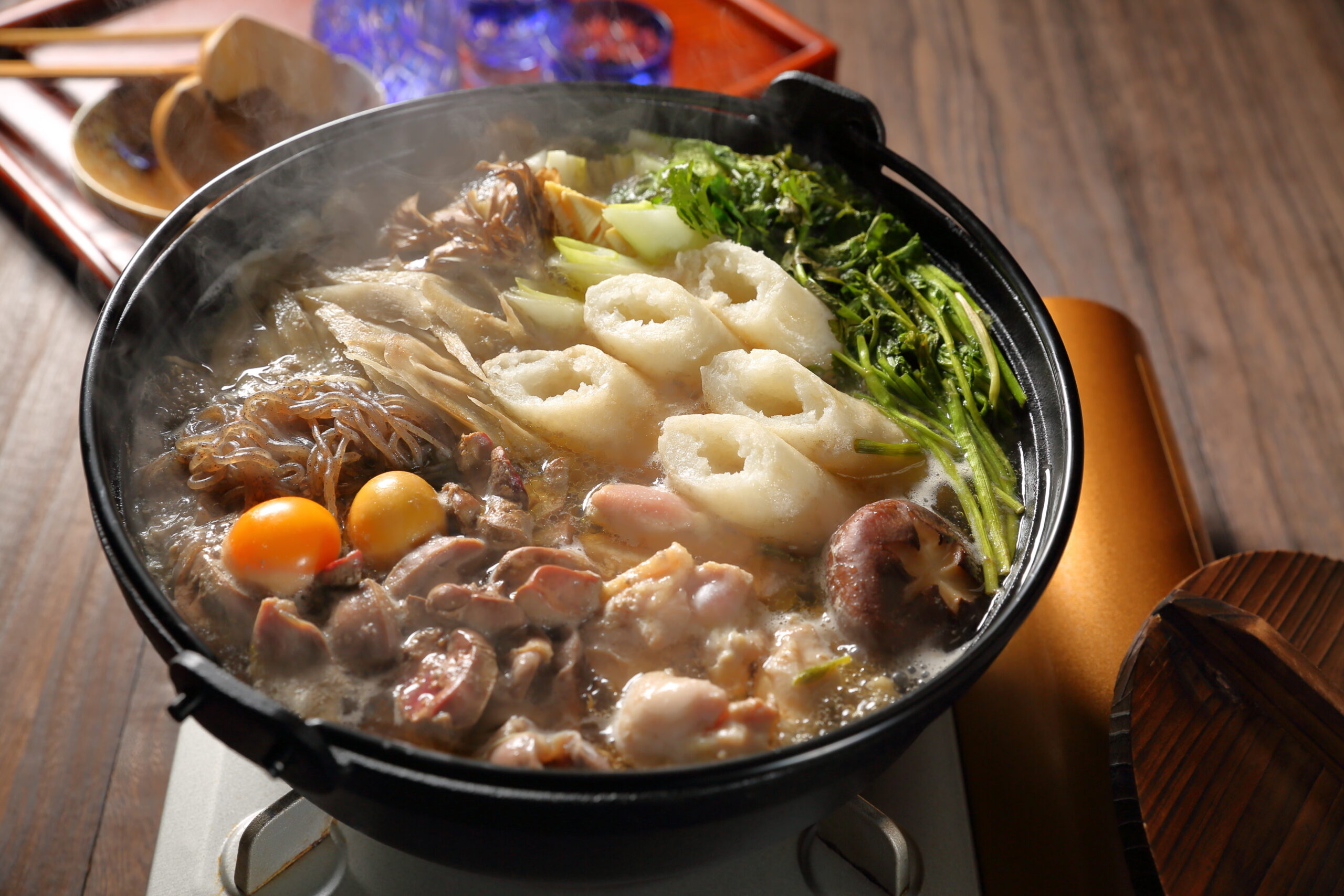 Steaming hot pot filled with meat, fish cakes, leafy greens, mushrooms, carrots, and glass noodles in broth on a portable stove.