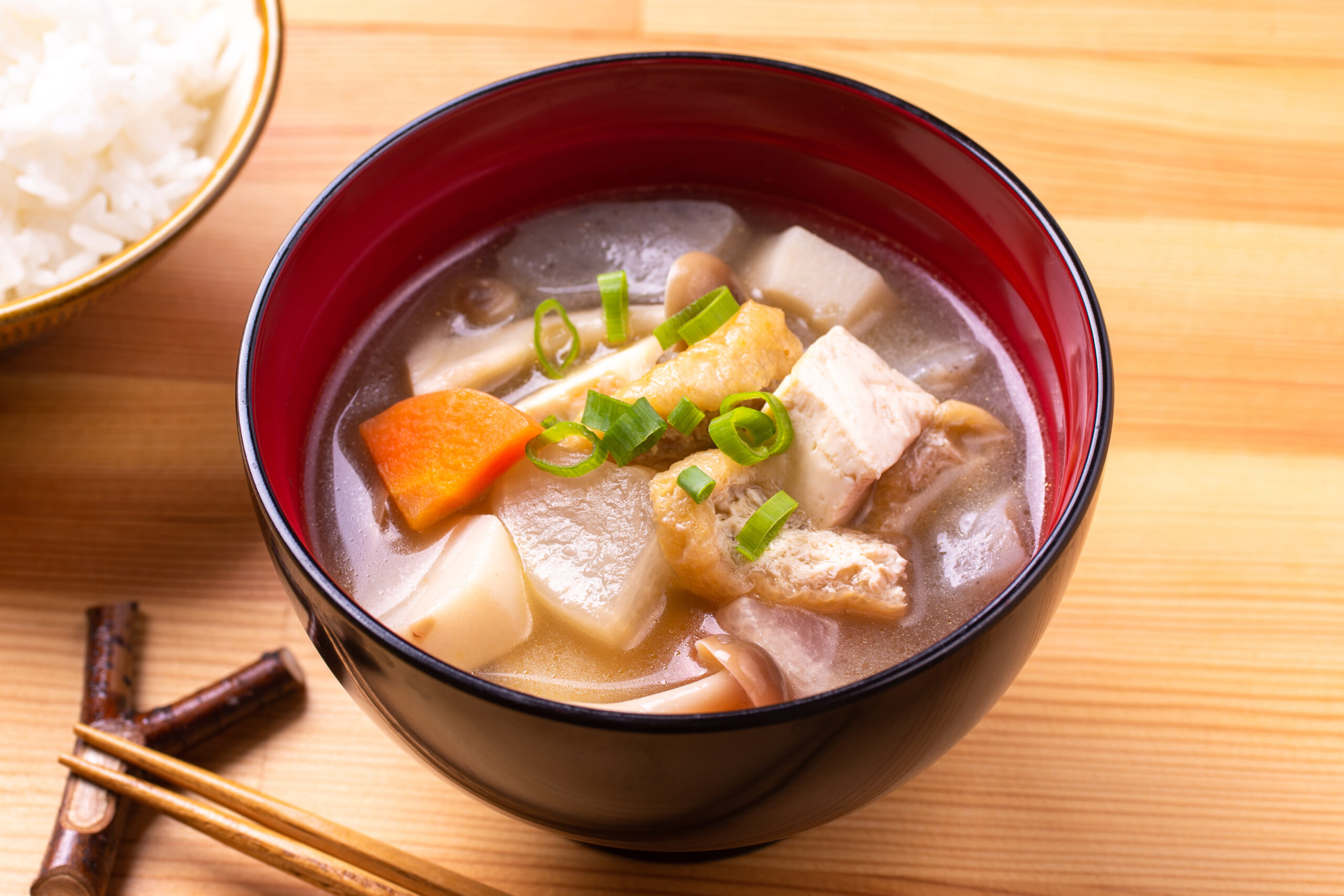 Bowl of miso-style soup with tofu, carrot, mushrooms, and green onions on a wooden table, with a side of rice. In the background is a separate rice bowl in the left edge.