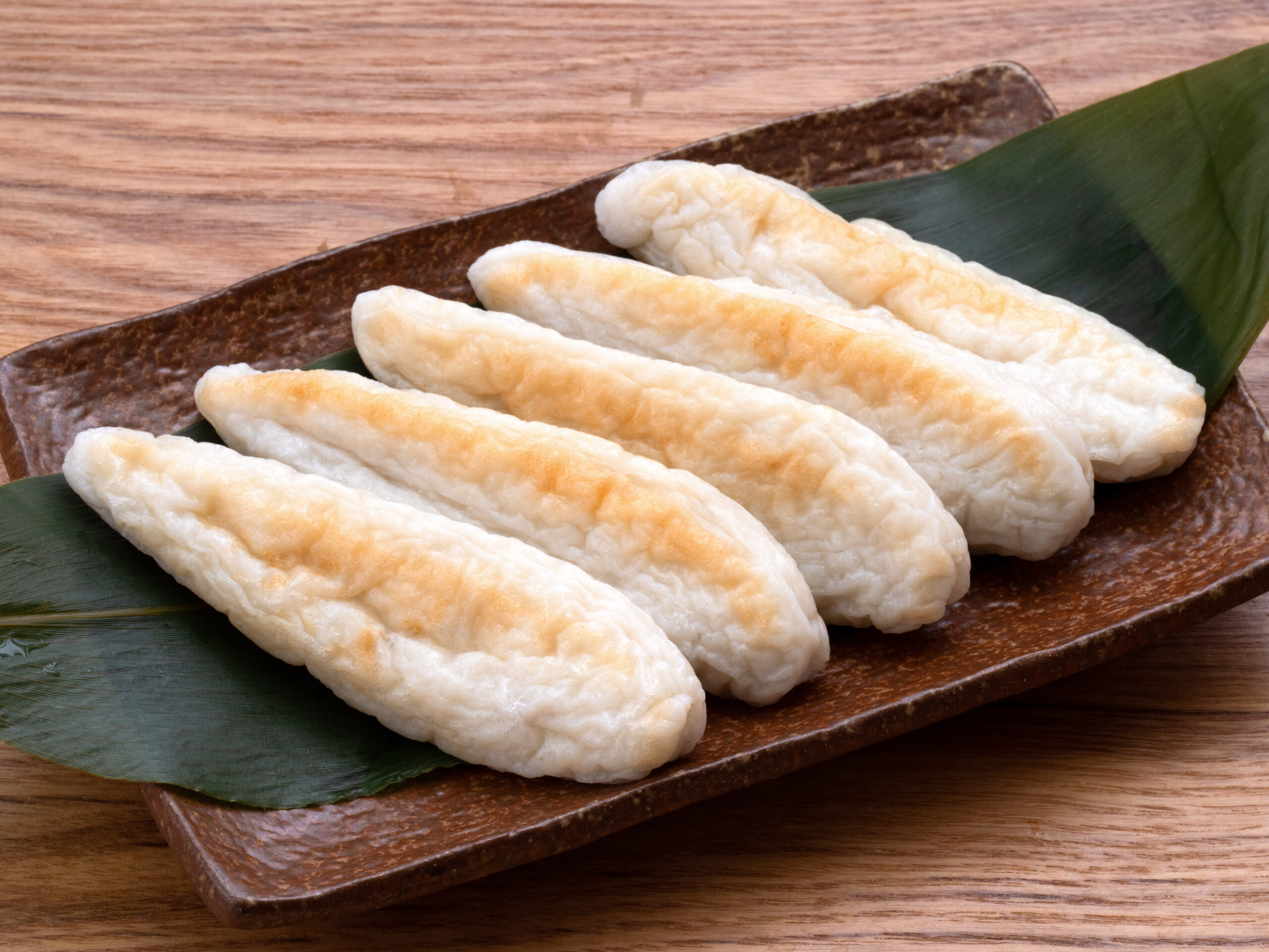 Rows of lightly browned white rice cakes (mochi) arranged on a brown ceramic platter with a green leaf garnish.