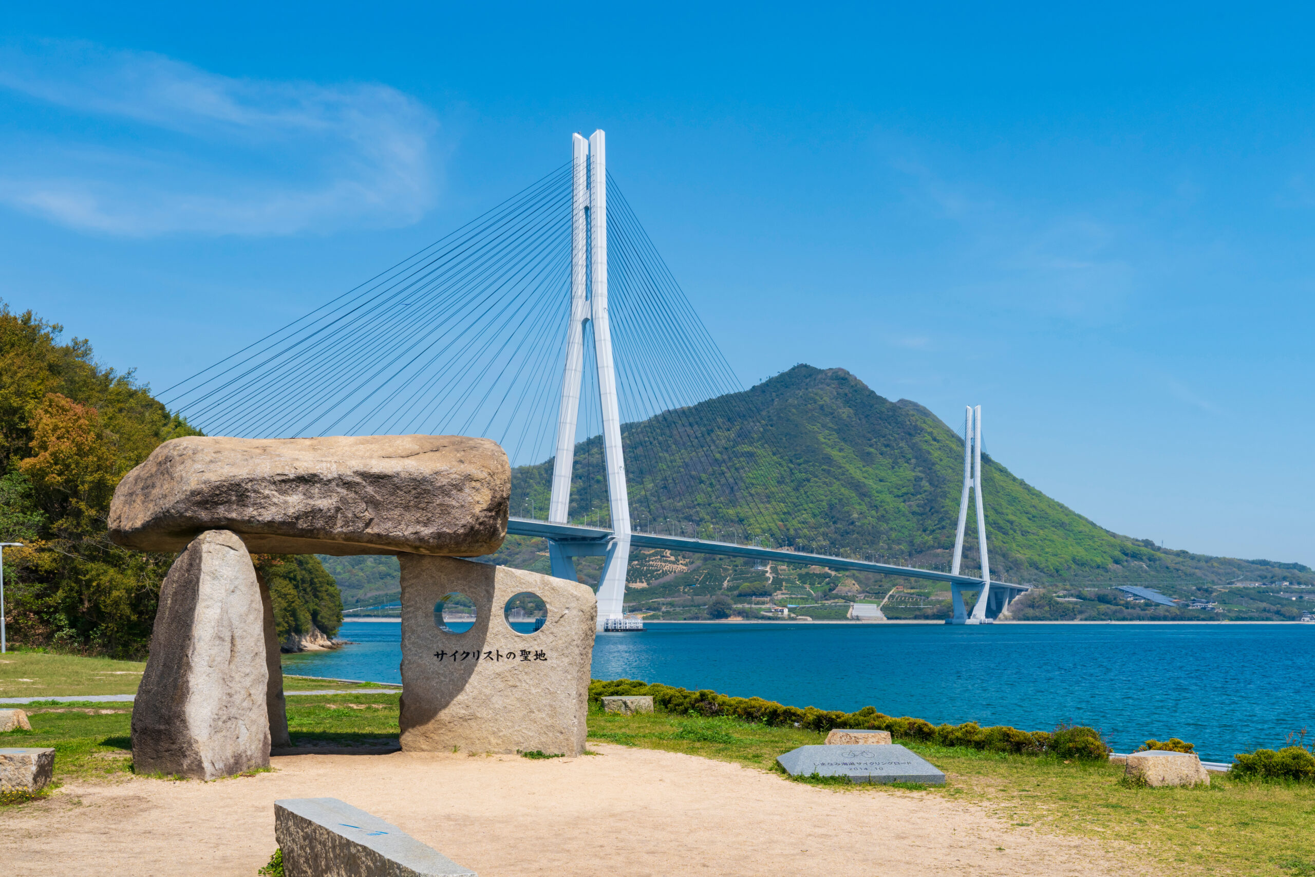 Cable-stayed bridge with two tall white towers and numerous cables over a blue lake, foreground rocks with Japanese inscription.