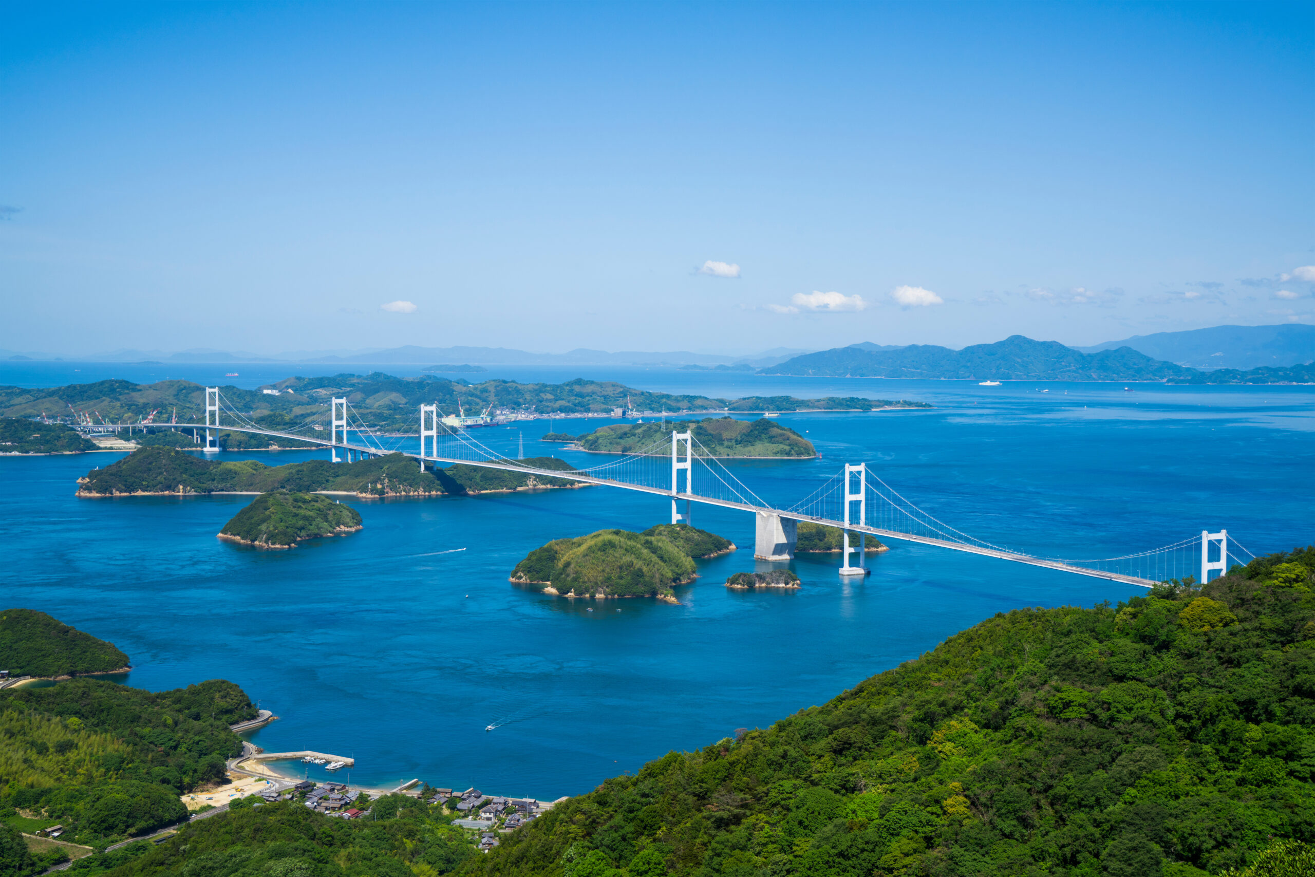 Suspension bridge crossing a blue bay with green islands and distant hills under a clear sky, aerial view