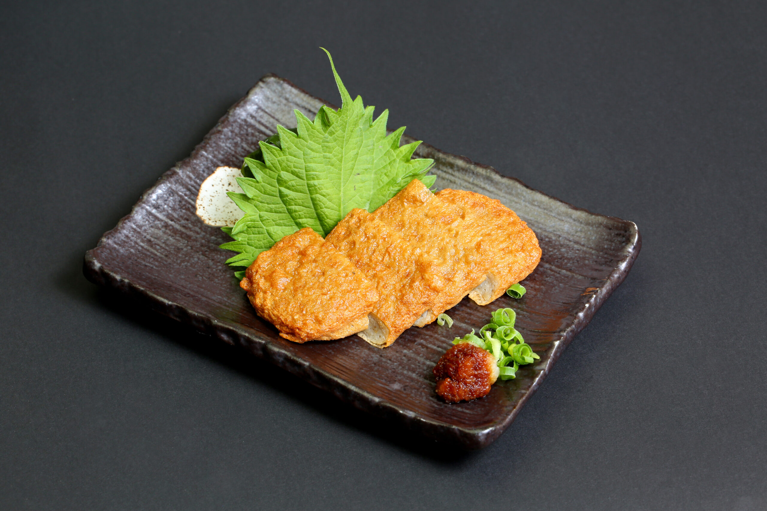 Golden fried fish fillet on a dark rectangular plate, garnished with a bright green shiso leaf and green onions.