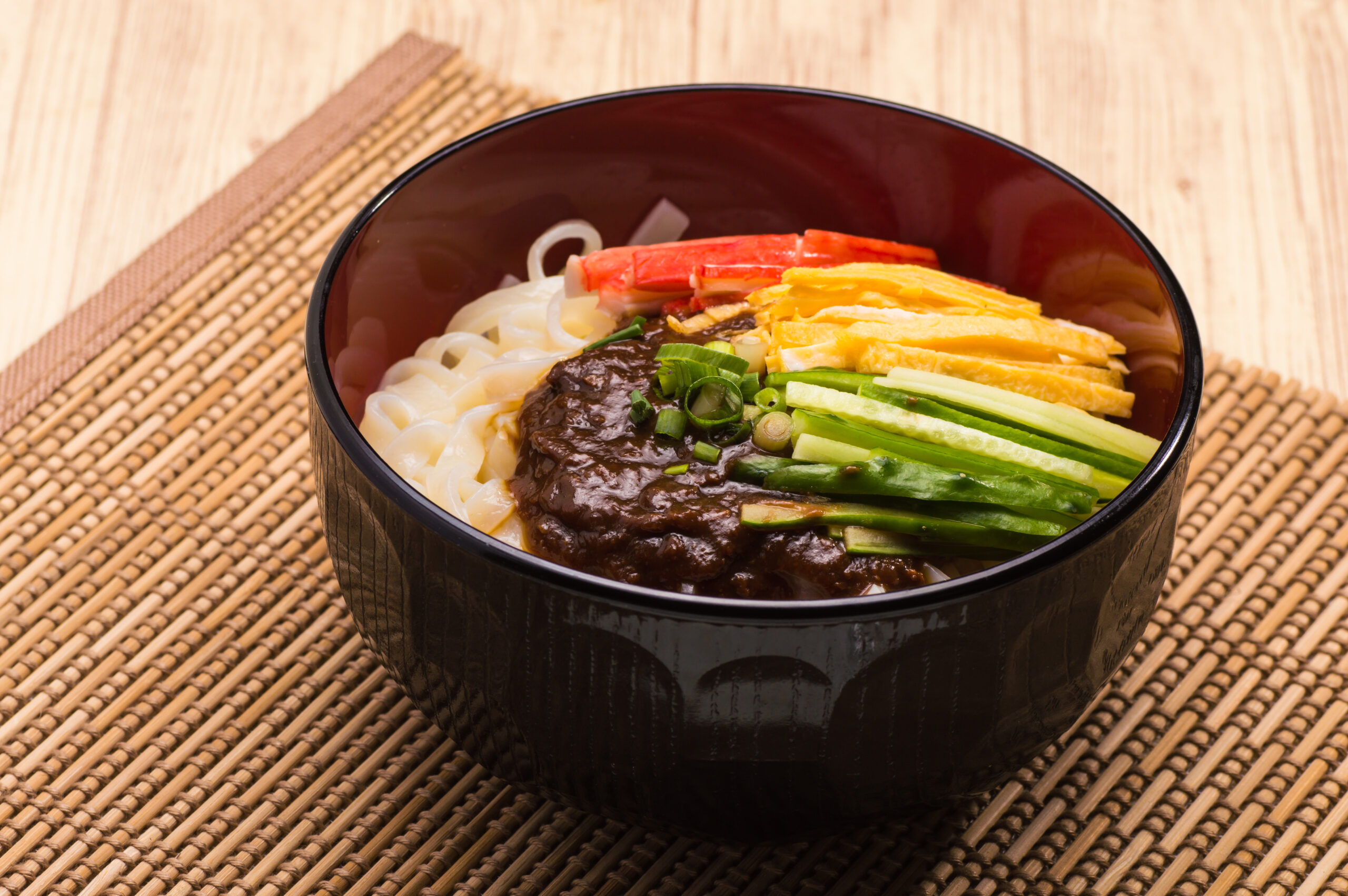 Udon noodles in a dark bowl with brown curry sauce, crab sticks, shredded egg, cucumber, and green onions on a woven mat.