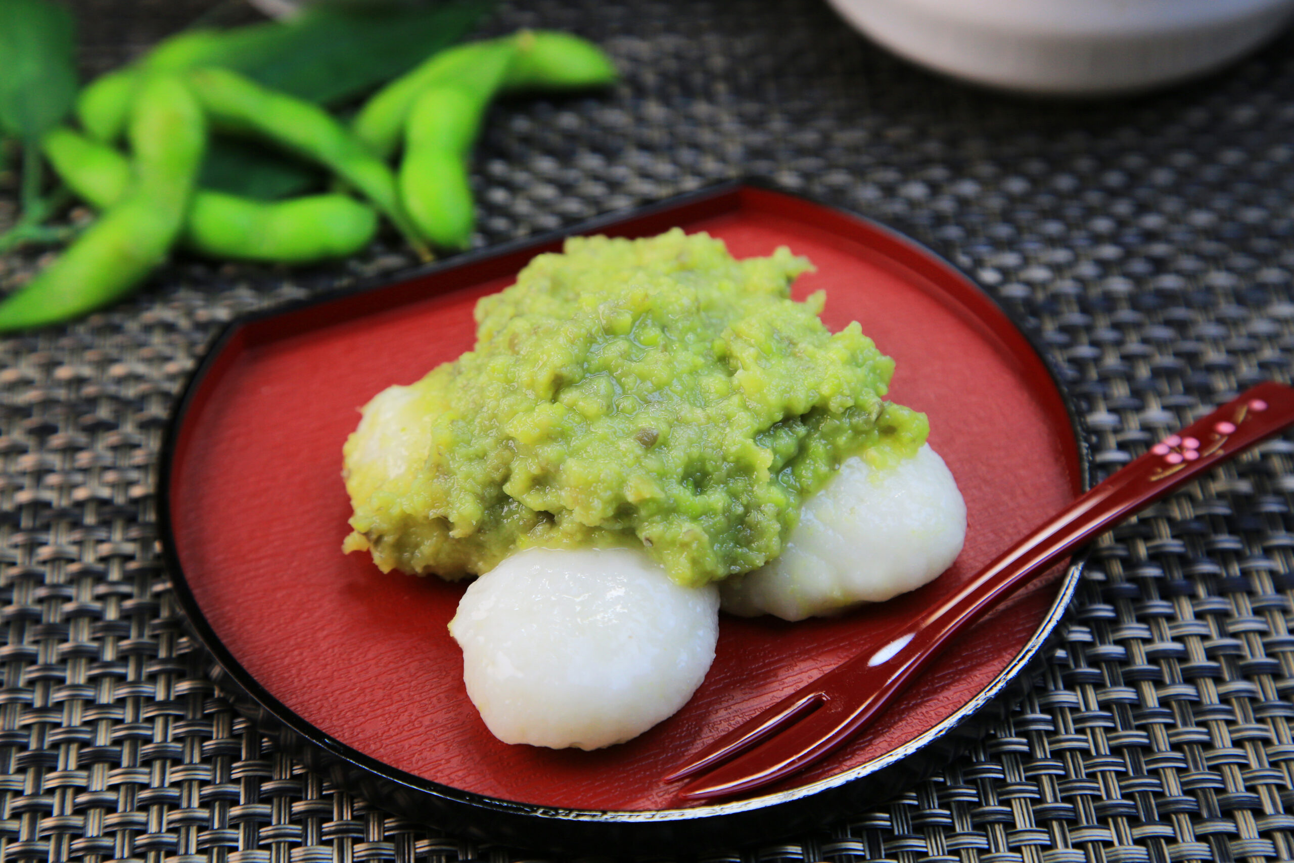 White mochi topped with a chunky green sauce on a red plate, with edamame in the background on a woven placemat.