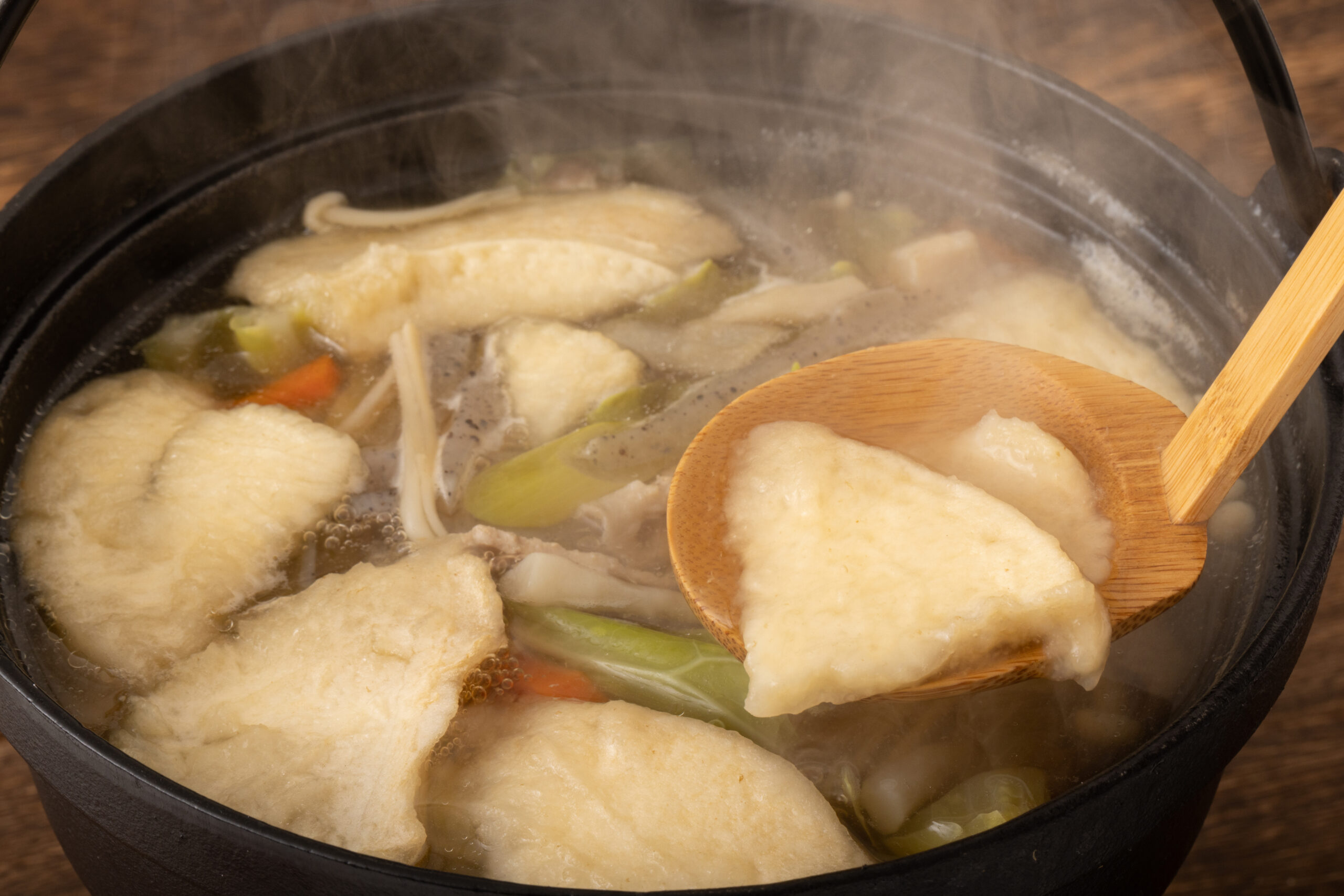 Simmering hot pot with tofu puffs, vegetables, and a wooden ladle resting inside, steam rising from the broth.