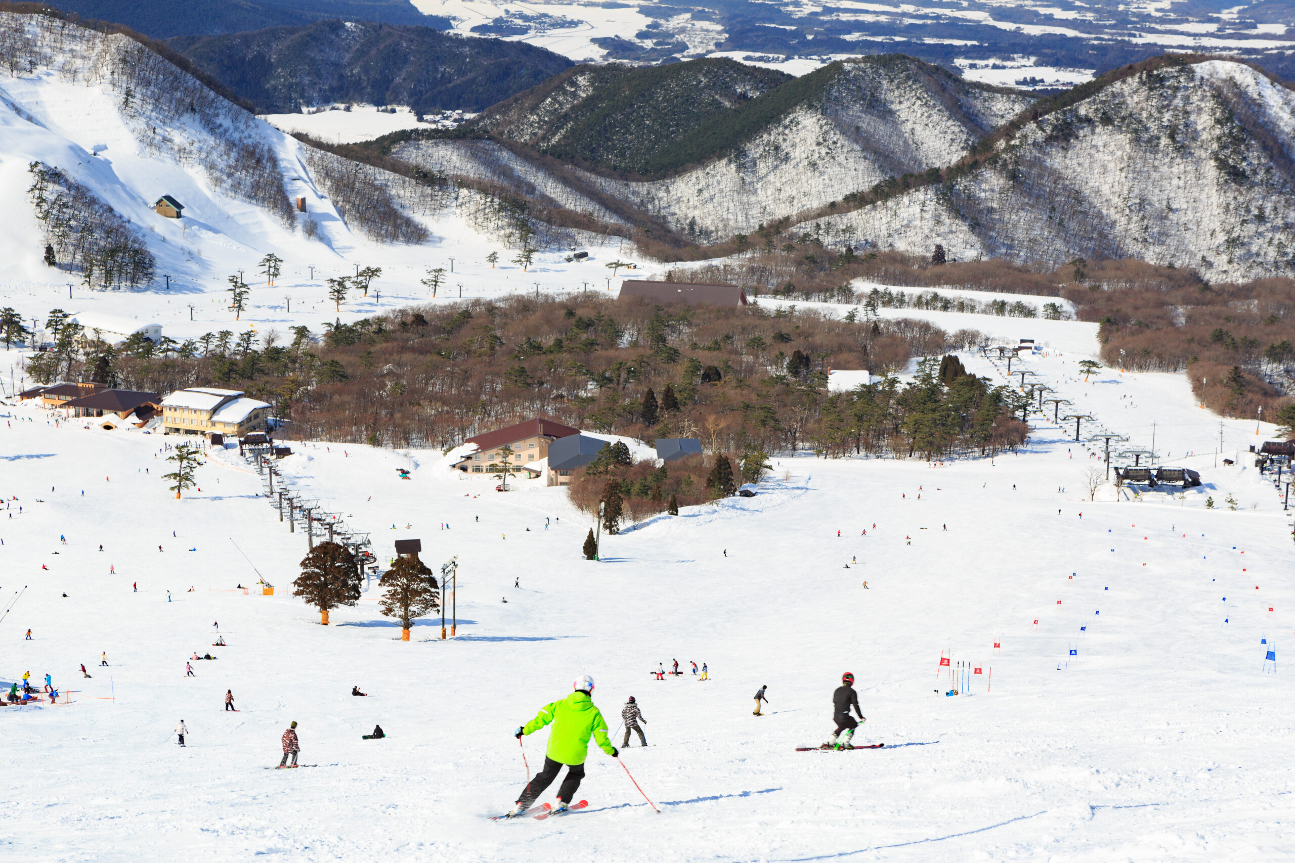 Snowy ski resort with skiers on a slope, chairlifts, and alpine buildings set against forested mountains.