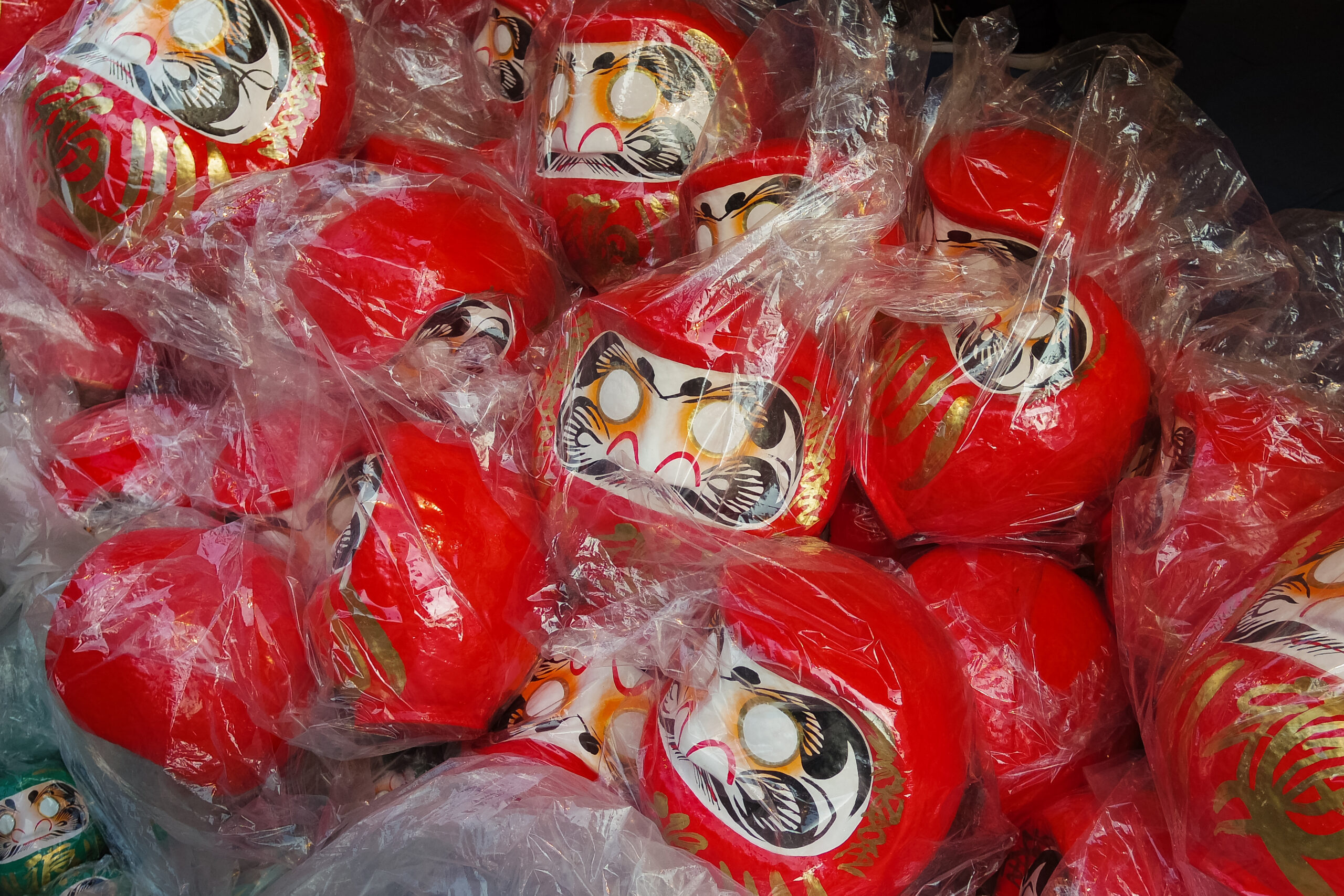 Red Daruma dolls with painted faces, wrapped in clear plastic bags, ready for display or sale in a shop window.