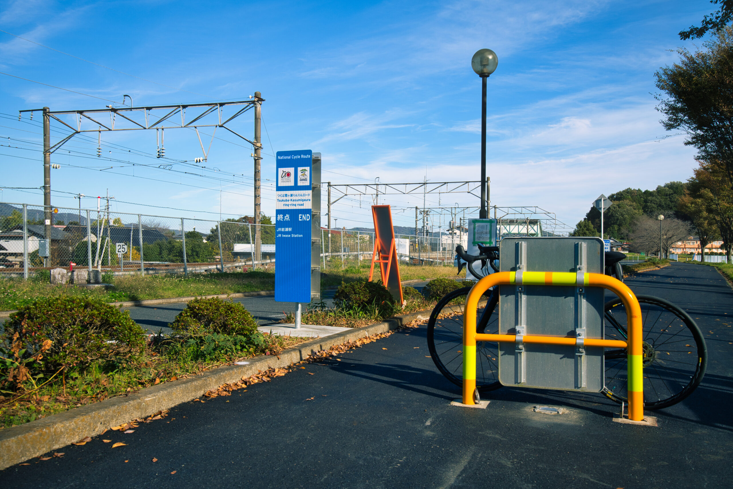 Railway area with overhead electric lines, a blue National Cycle Route sign, and a bright orange barrier on a sunny day close to a bike rack/upright bike.