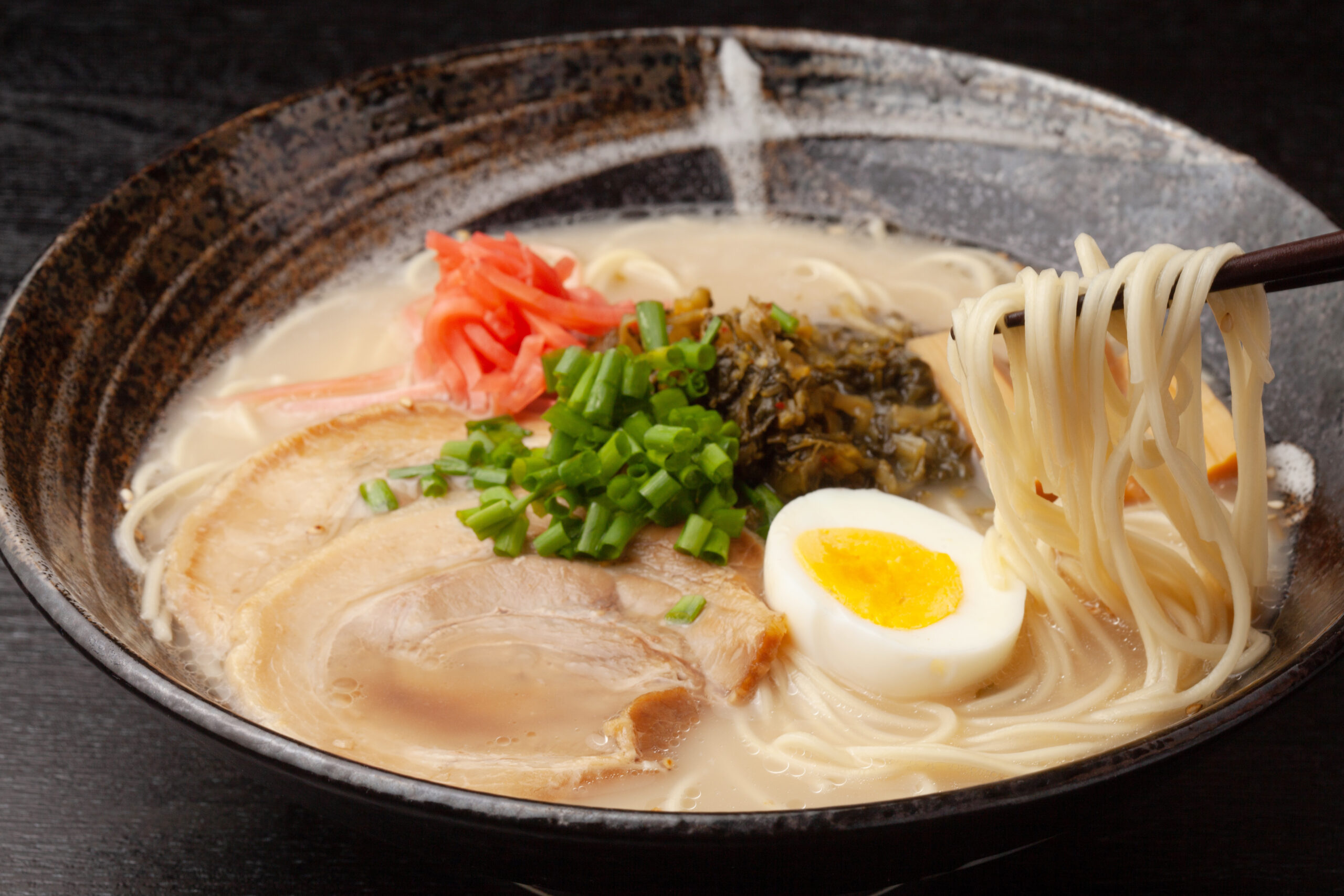 Bowl of ramen with creamy broth, sliced pork, halved soft-boiled egg, green onions, pickled ginger, and noodles visible.
